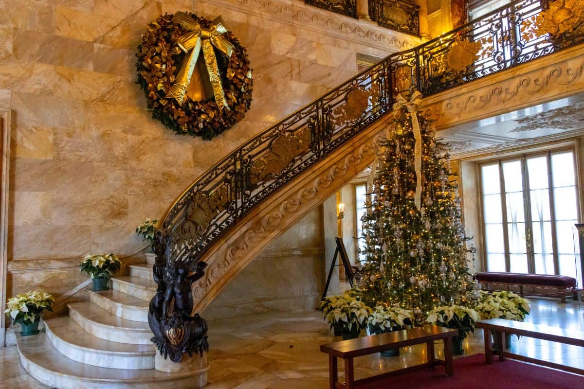 A decorated marble staircase with a large wreath on the wall and a Christmas tree with gold ornaments beside it. White poinsettias are arranged along the steps and tree base.