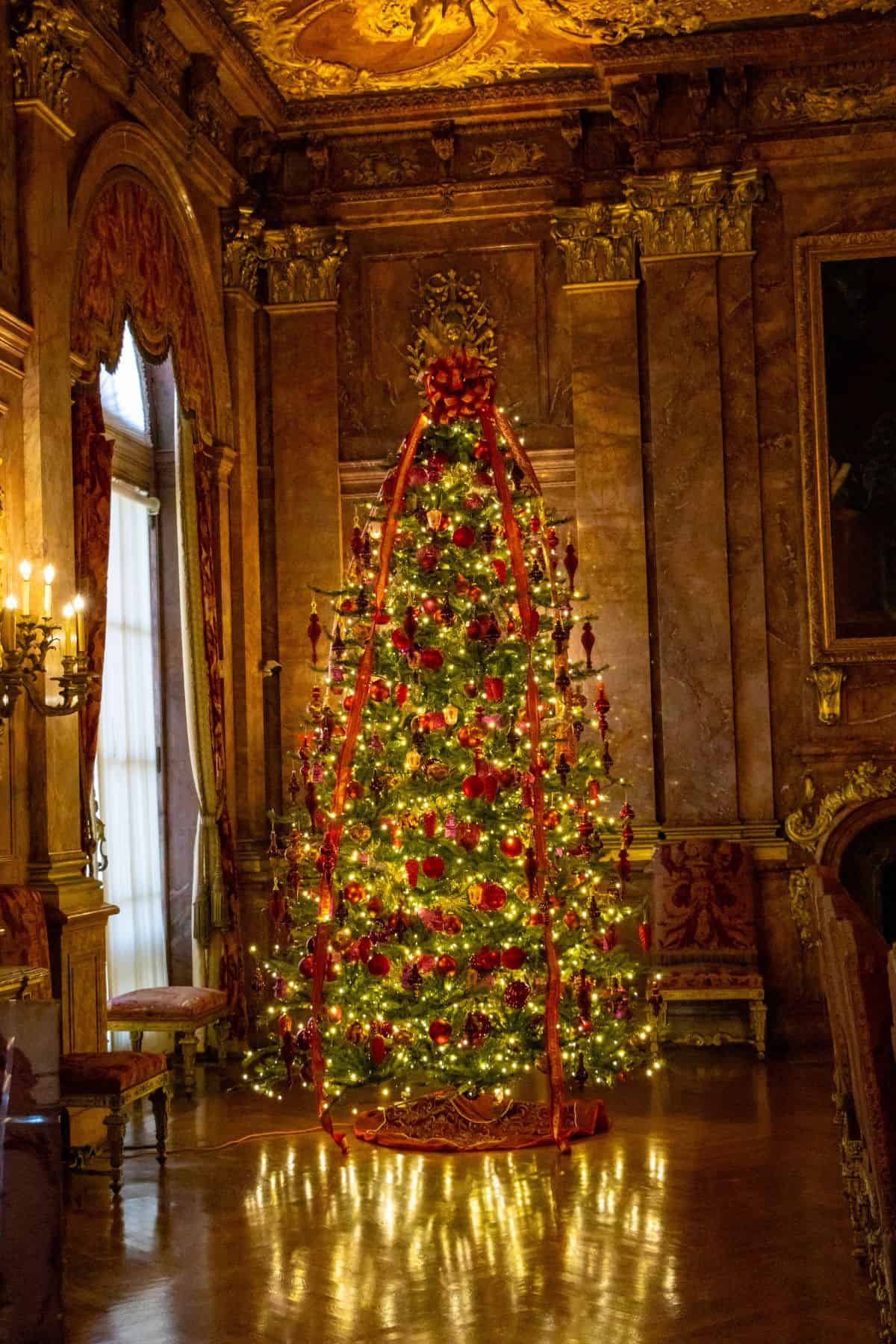 A Christmas tree decorated with red ornaments and ribbons stands in an ornate, warmly lit room with tall windows, gold accents, and detailed wall paneling.