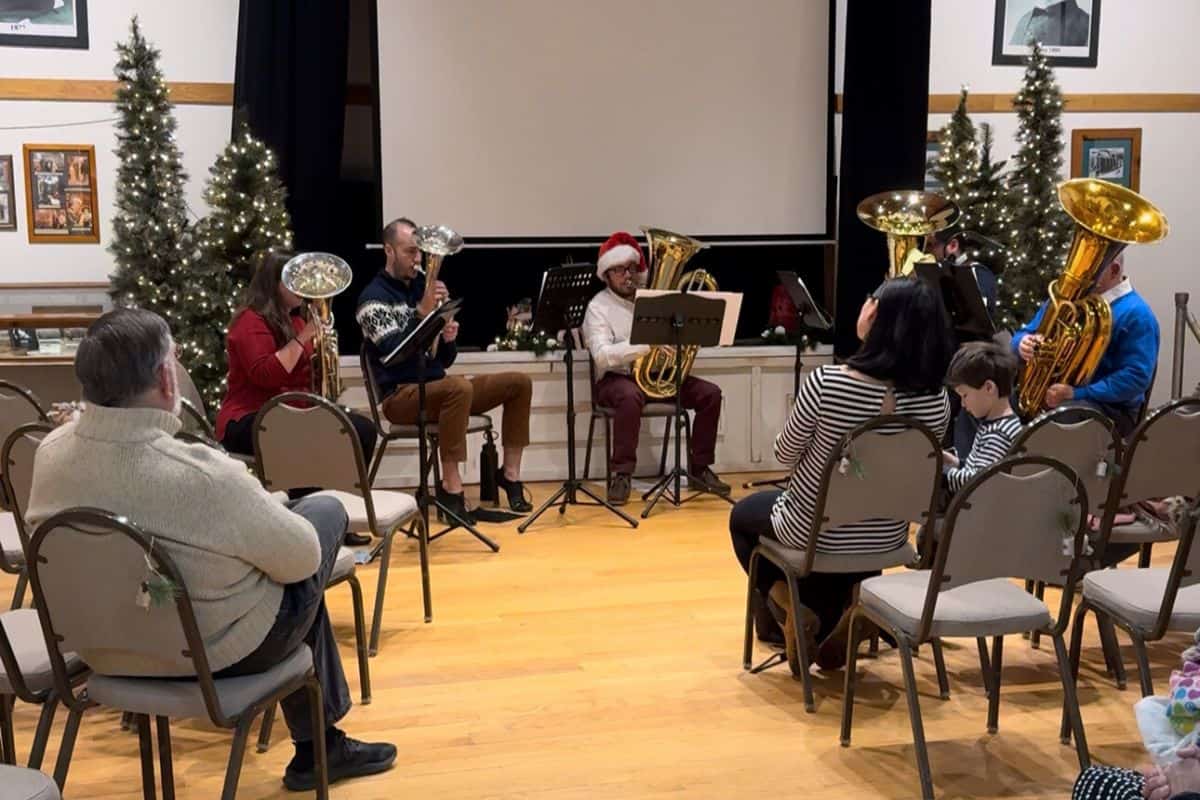 A small brass band performs in front of an audience in a decorated room with Christmas trees and framed photos on the walls.