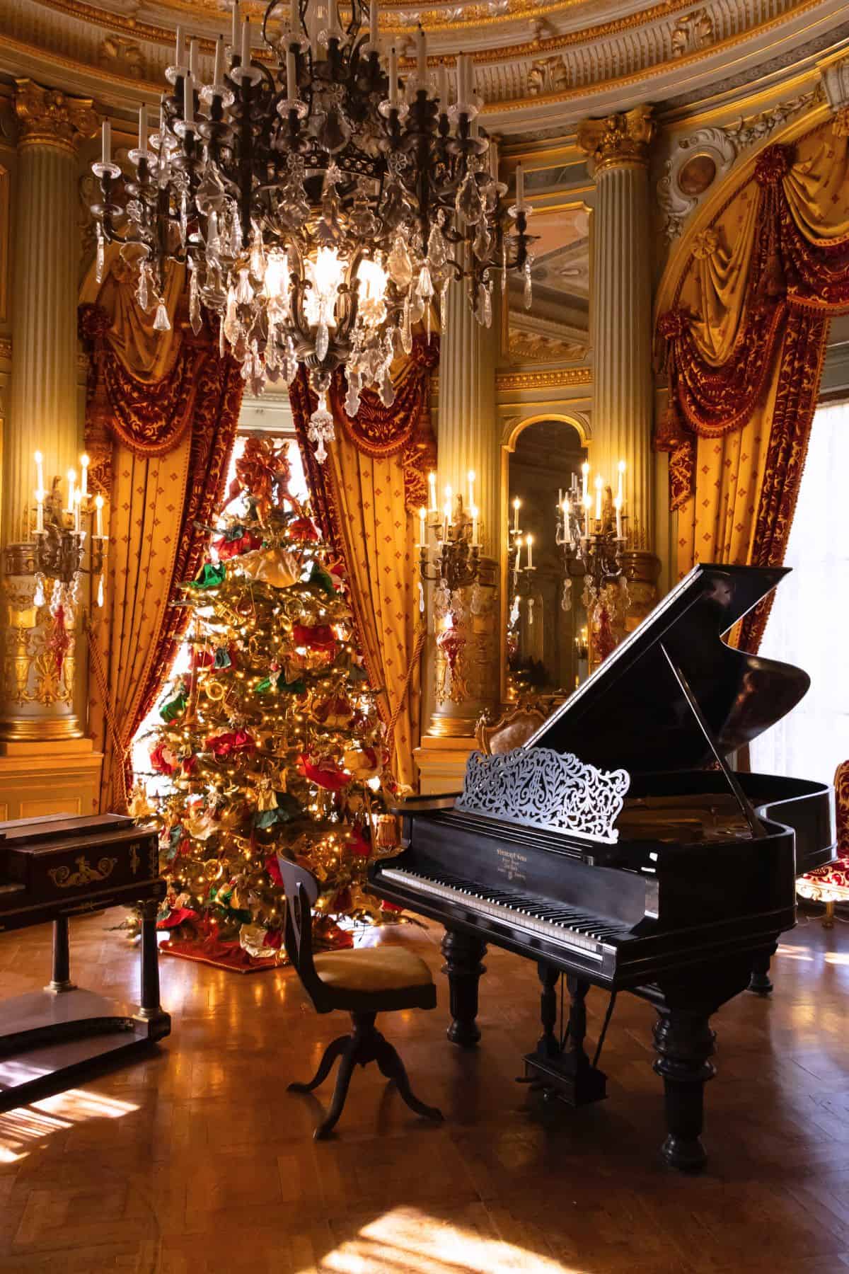 A grand piano and chair sit in an ornate room with a decorated Christmas tree, chandeliers, tall columns, and gold drapery.