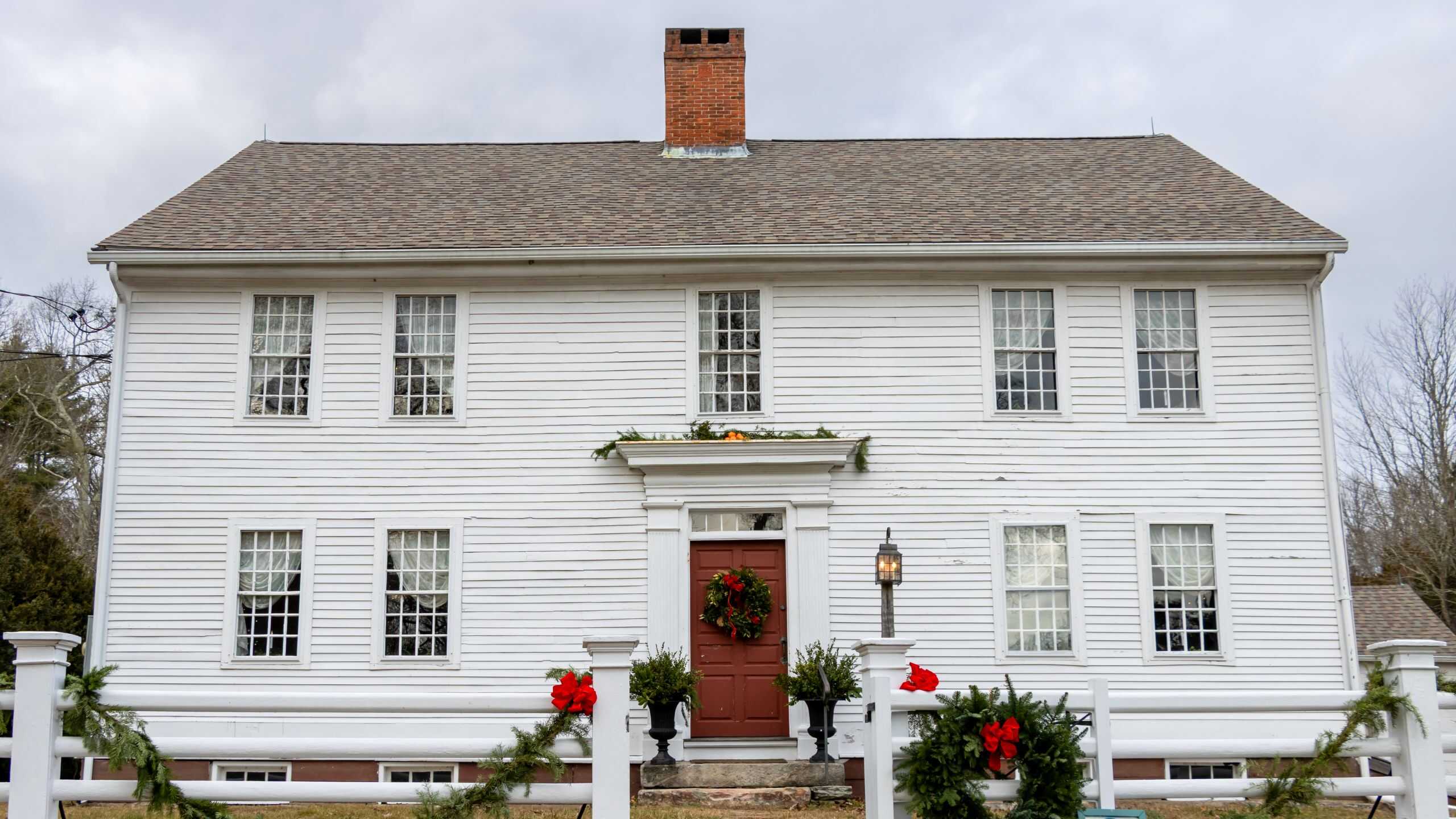 A two-story white wooden house with a central red door, holiday wreaths, and garlands on the fence and above the entrance under a cloudy sky—perfect inspiration for festive things to do in Connecticut this holiday season.
