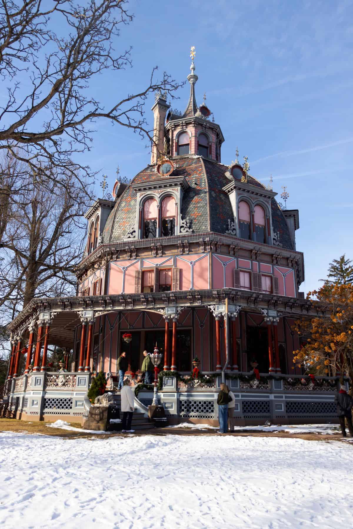 A large, ornate Victorian house with a round tower, elaborate trim, and red accents, surrounded by snow and leafless trees under a clear blue sky.