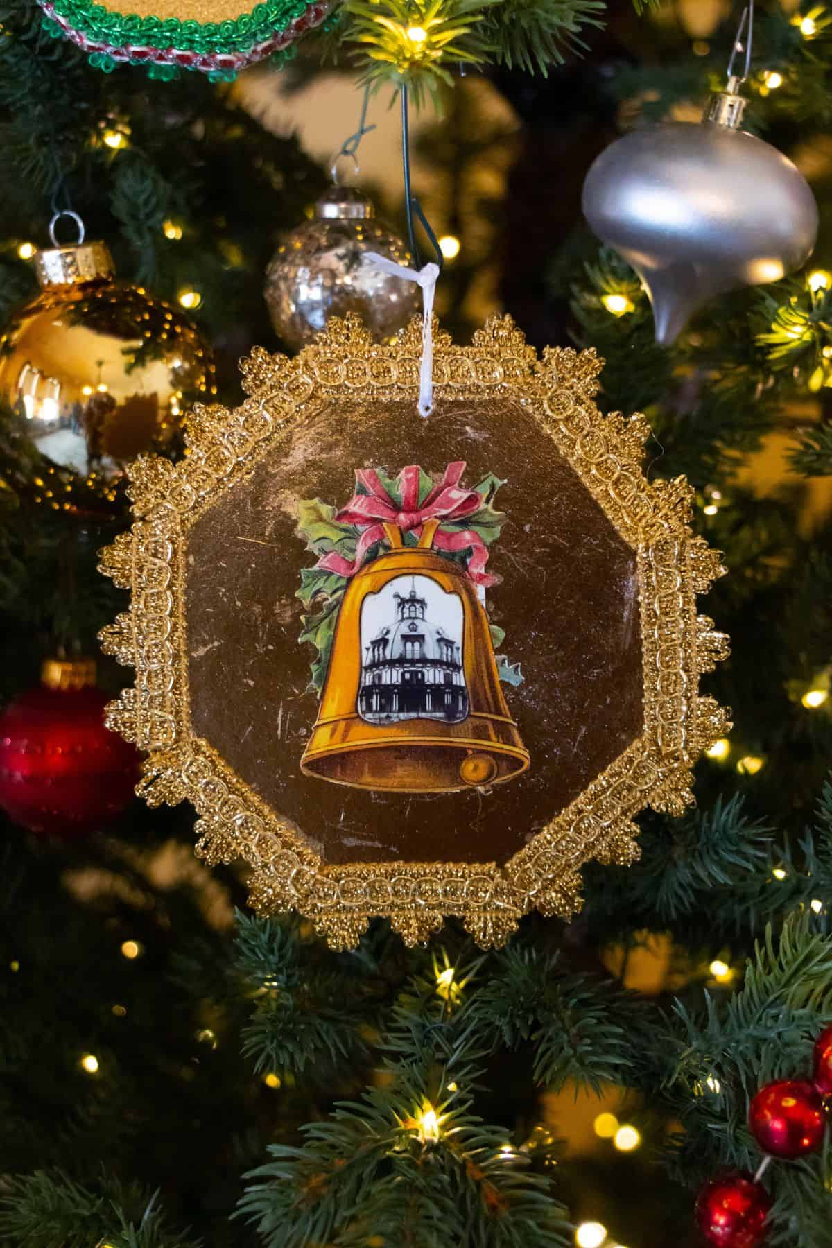 Gold and brown octagonal Christmas ornament with an illustration of a bell and building, hanging on a decorated tree with lights and other ornaments.