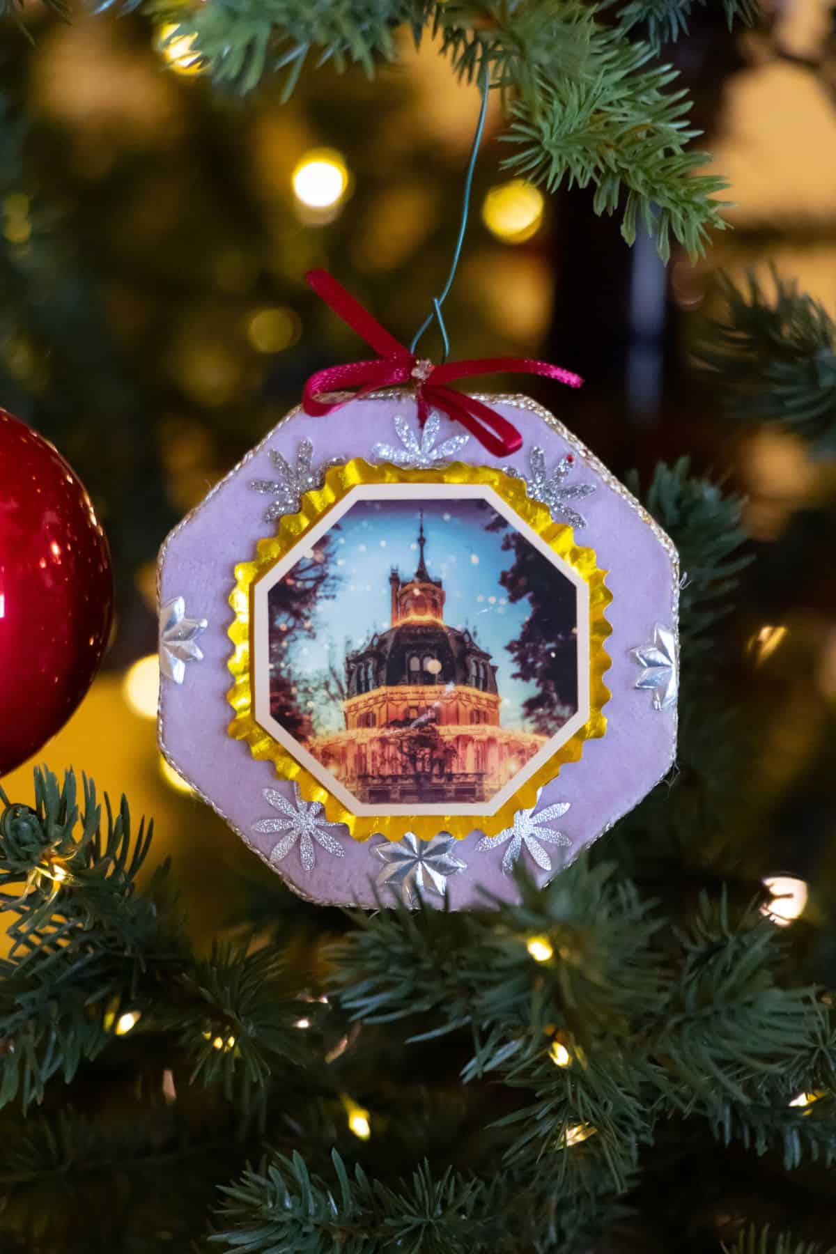 A Christmas tree ornament featuring a photo of a lit-up clock tower, decorated with a red bow, silver stars, and gold trim, hangs on a tree branch next to a red bauble.