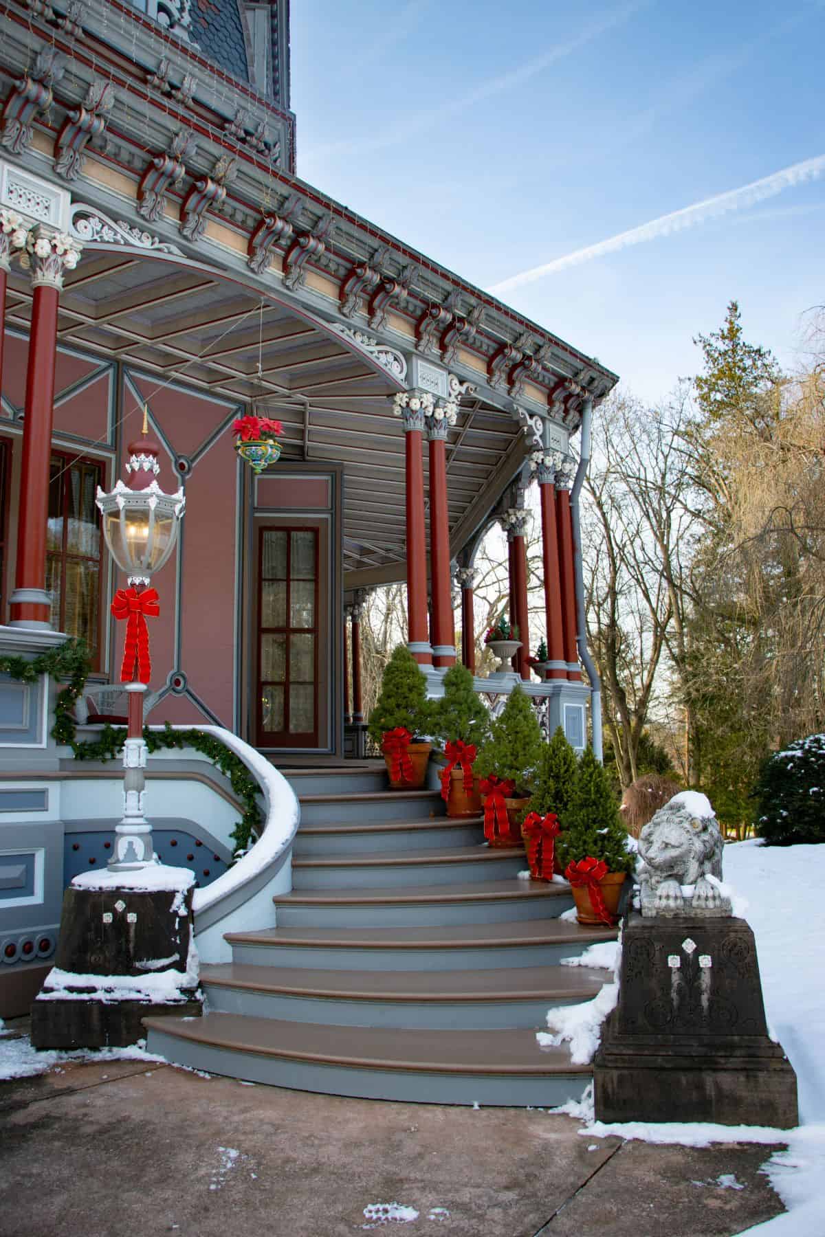Victorian-style house with ornate porch, decorated with red bows and potted plants, snowy ground, stone lion statue by the steps.