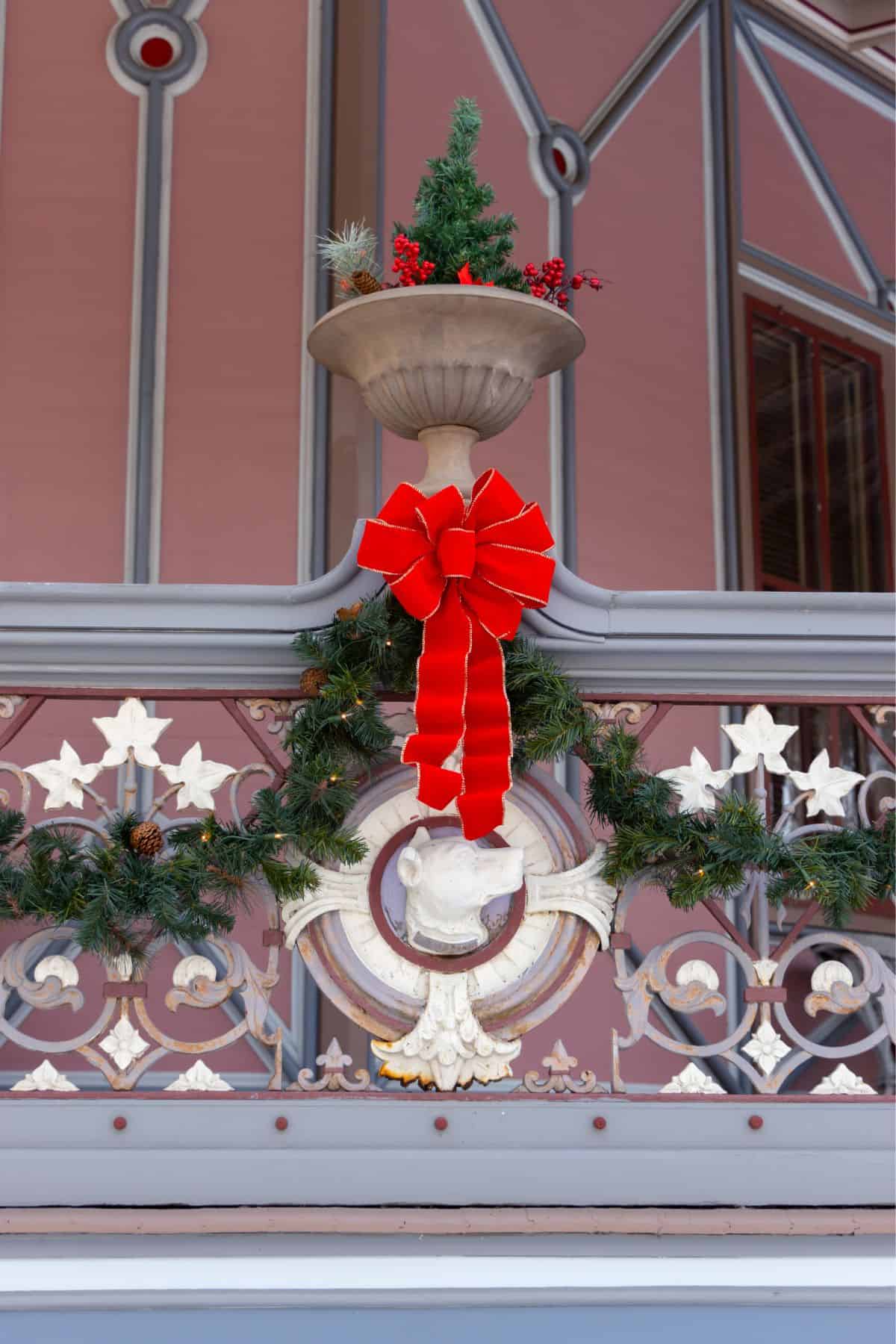 Festive garland and a bright red bow decorate a metal railing with a sculpted fish, topped by a planter with a small artificial tree and berries on a pink building.