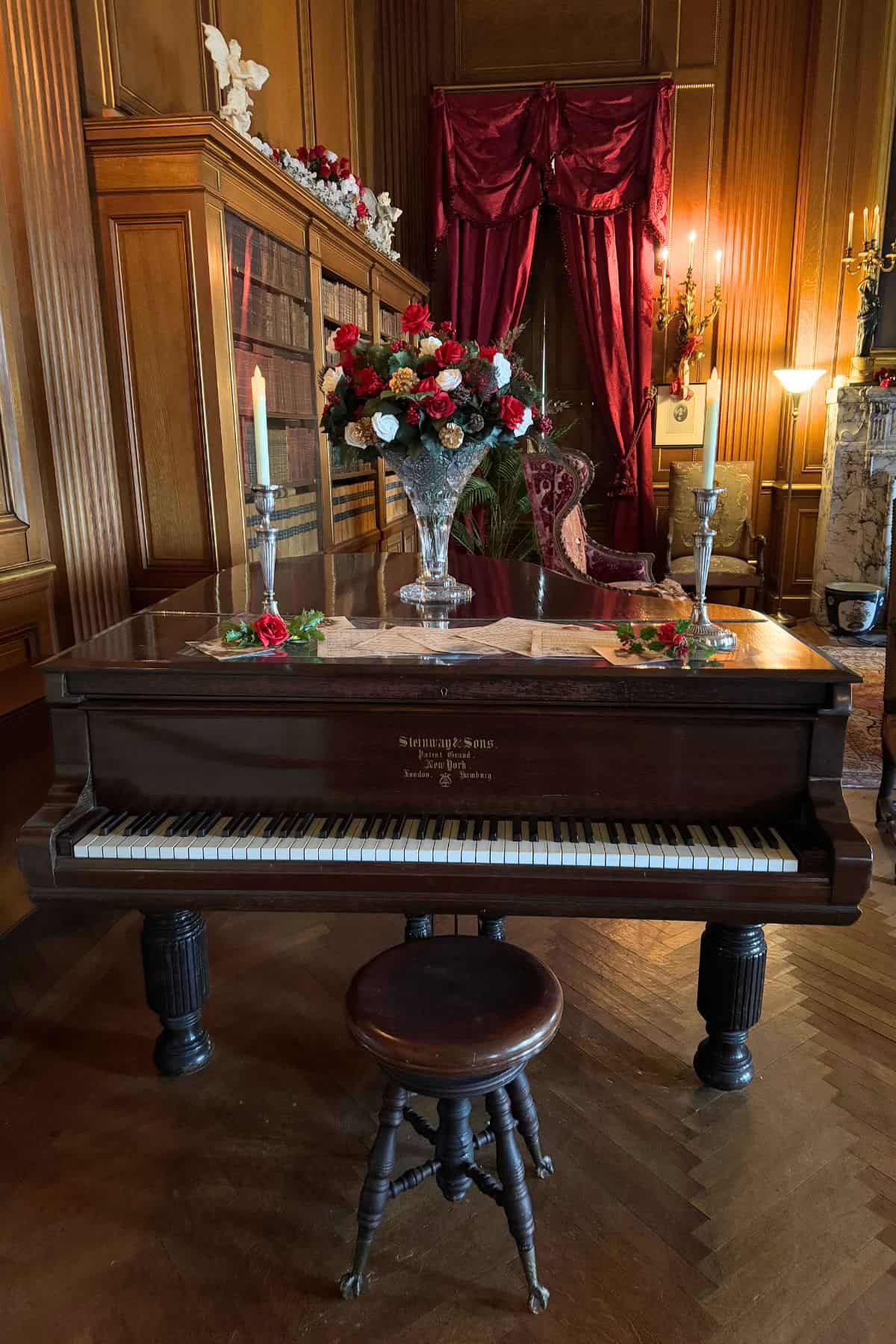 A grand piano with floral arrangements and candles sits in an ornate room with wood paneling, red drapes, and antique furniture.