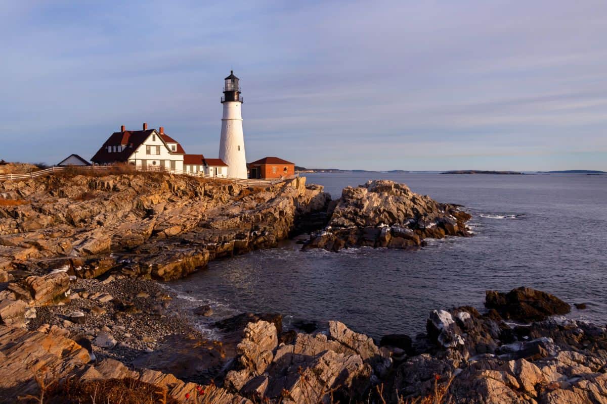 A white lighthouse with an attached house sits on a rocky coastline overlooking calm ocean waters under a partly cloudy sky.