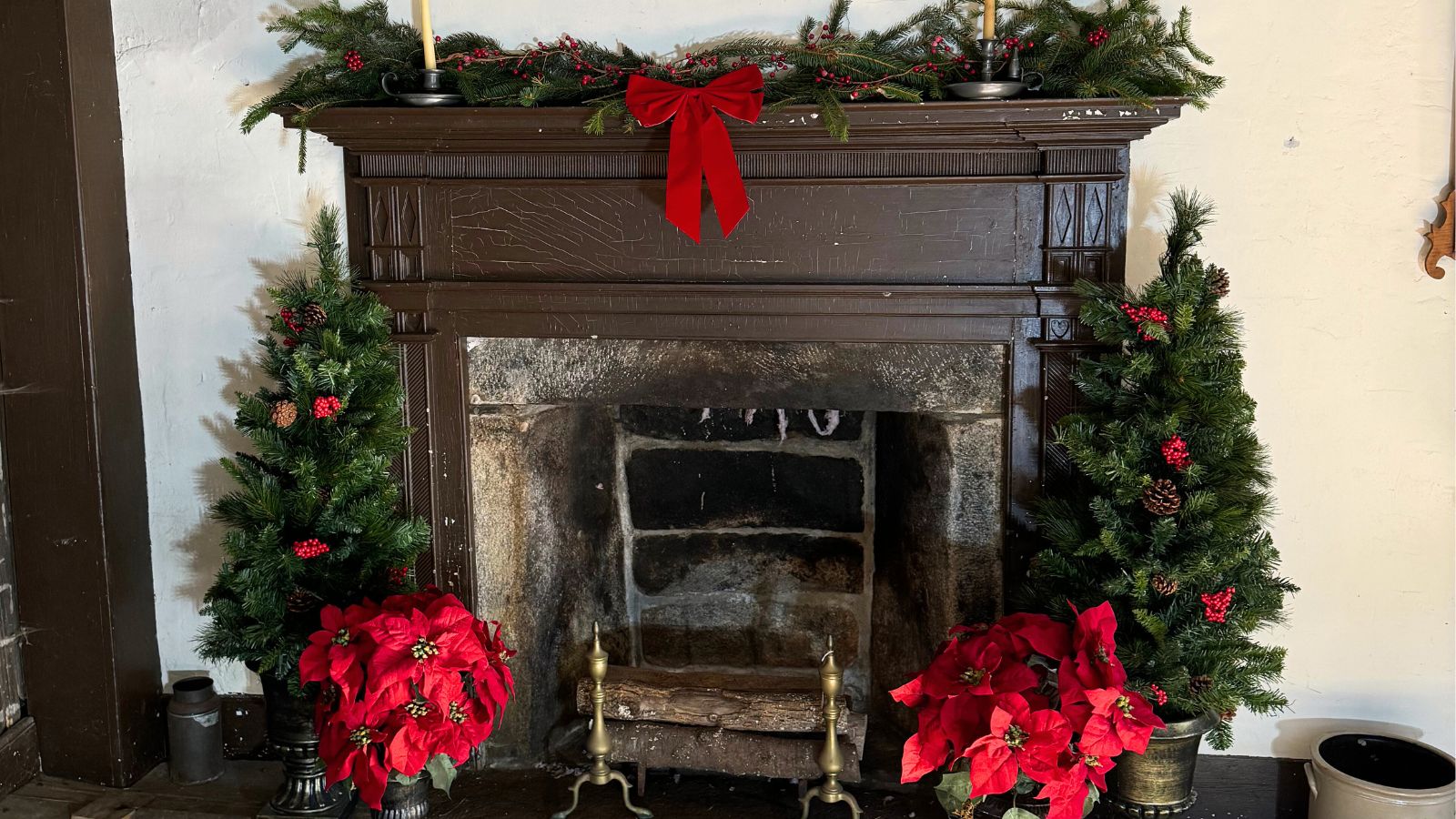 A decorated fireplace with a red bow, garland, two small Christmas trees, poinsettias, and brass andirons on the hearth.