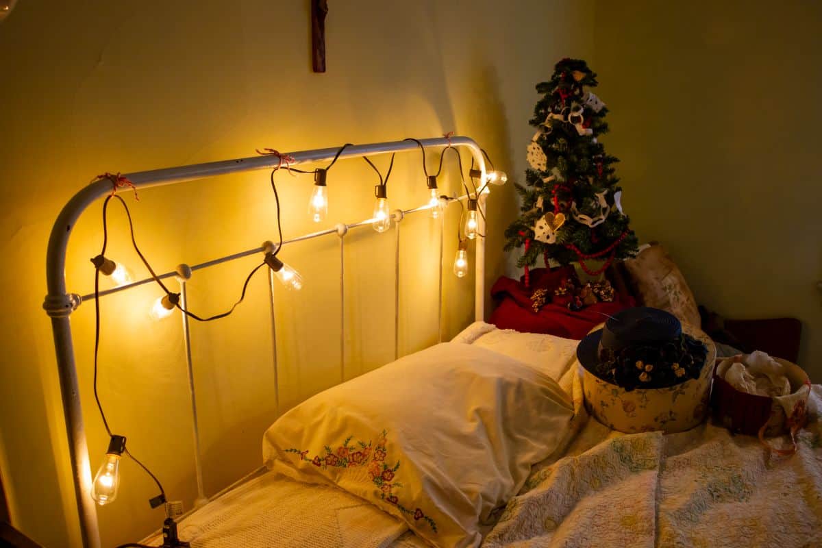 A bed with a white metal headboard decorated with string lights, next to a small decorated Christmas tree and storage boxes on a quilted bedspread.