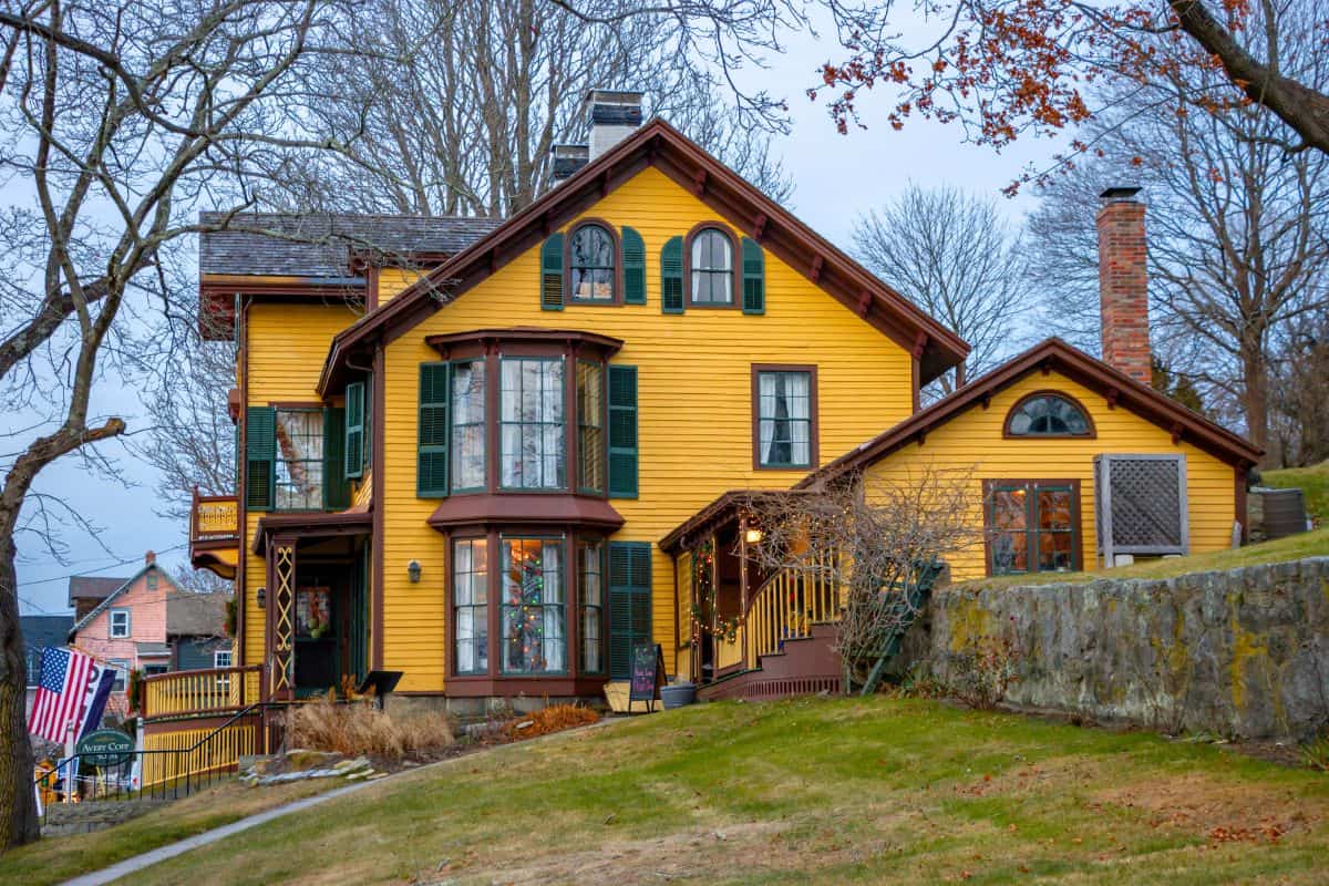 Large yellow house with green shutters, brown trim, and multiple gabled roofs, set on a sloped lawn with bare trees and a stone retaining wall in the foreground.
