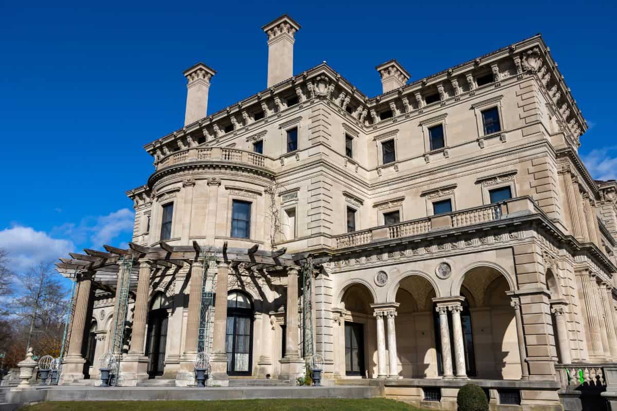 A large, ornate stone mansion with columns, arches, and multiple chimneys stands under a clear blue sky.