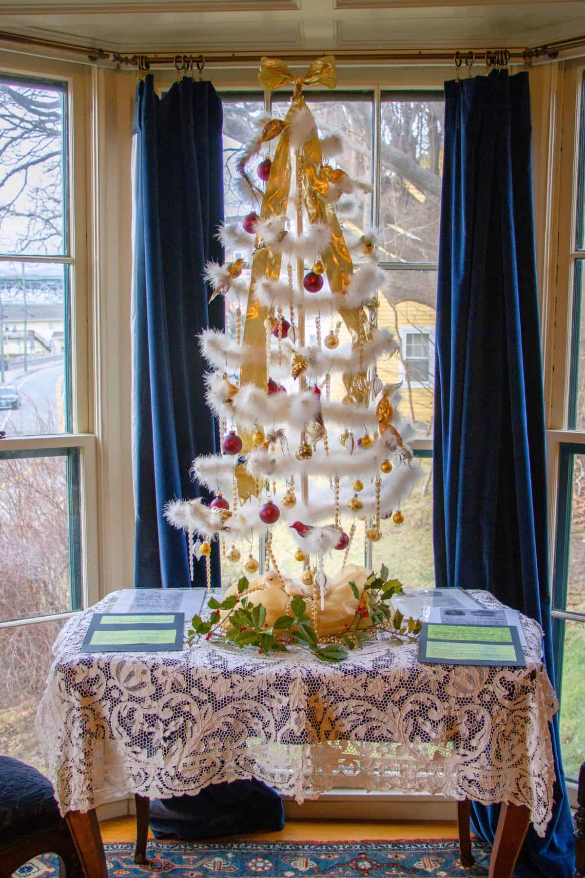 White artificial Christmas tree decorated with gold ribbons and red ornaments, displayed on a lace-covered table in front of large windows with navy blue curtains.