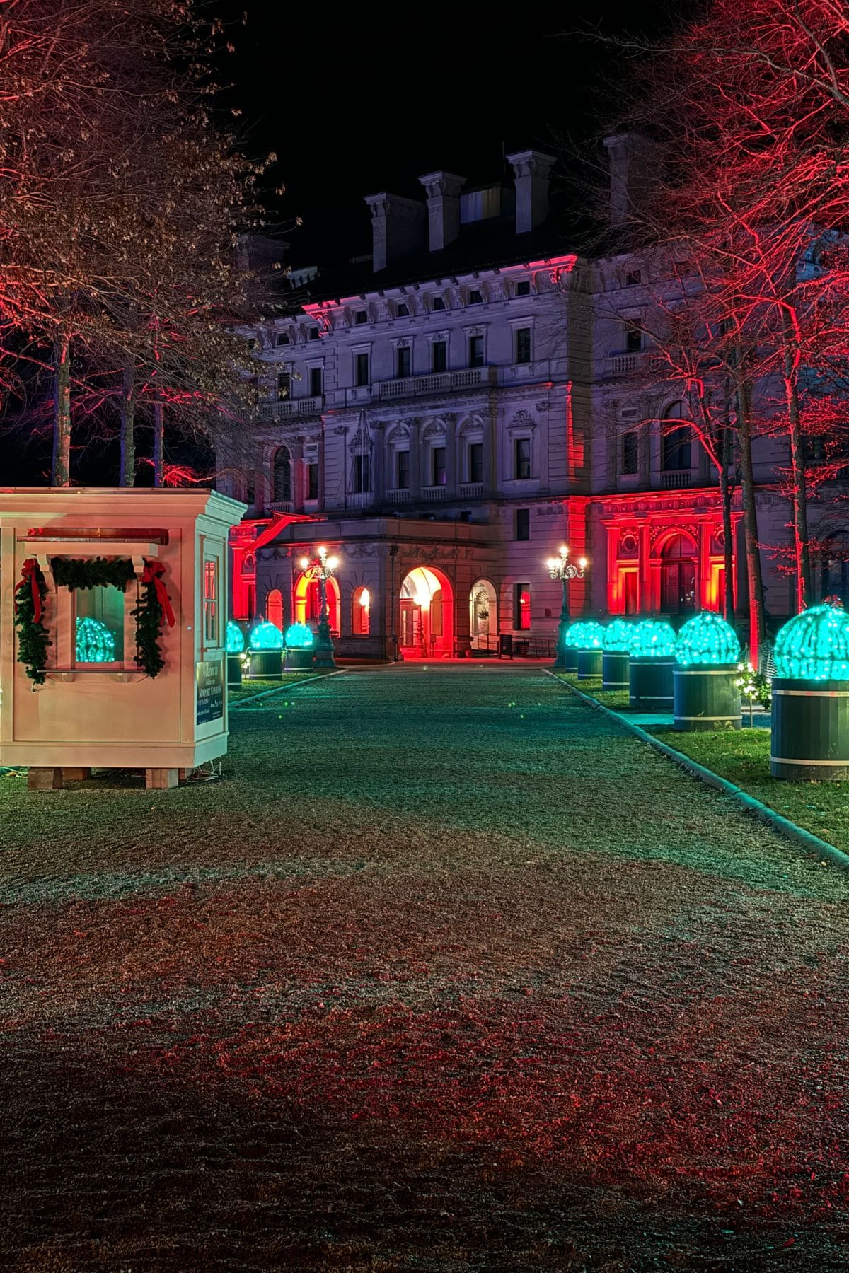 A large building glows with red lights at night during Holidays at the Newport Mansions, featuring illuminated dome-shaped decorations and bare trees along a gravel path. A small white hut trimmed with garland sits to the left.