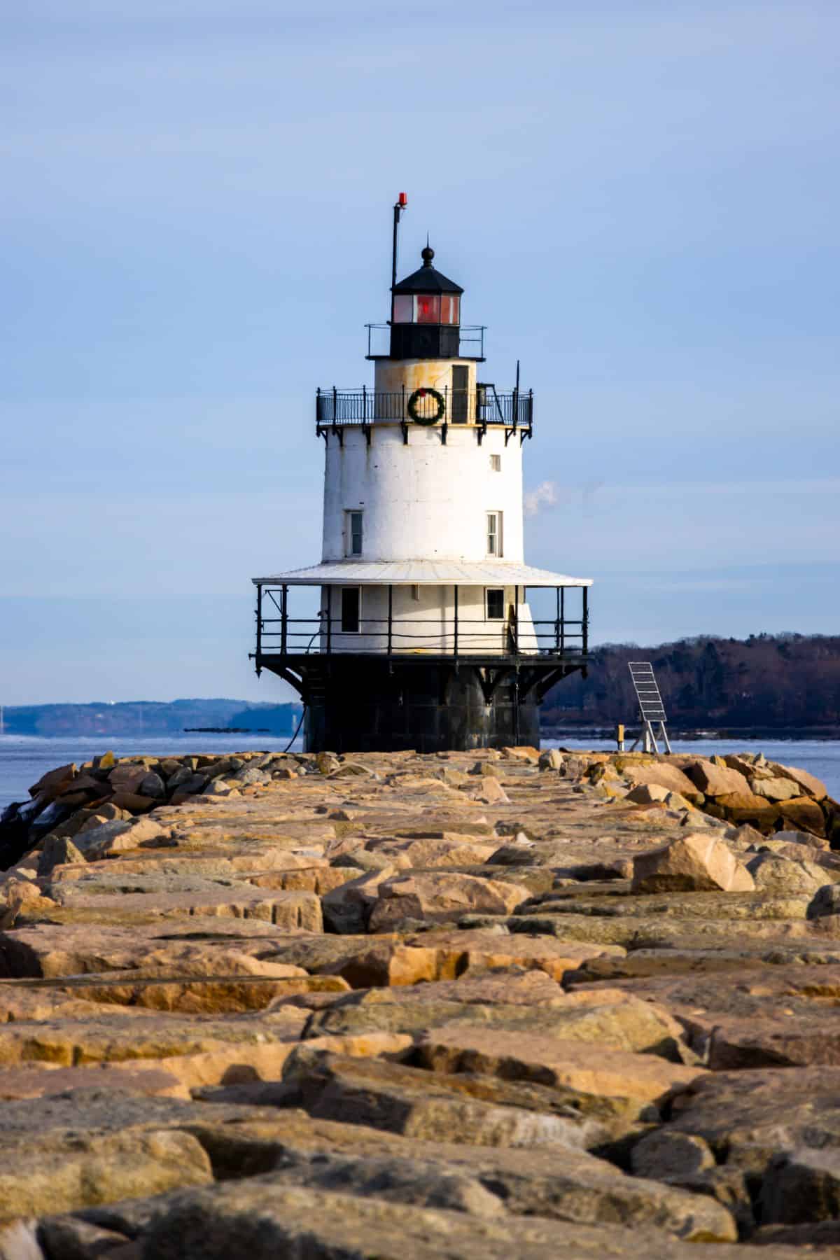 A black and white lighthouse stands at the end of a rocky pier, with calm water and a distant shoreline in the background.