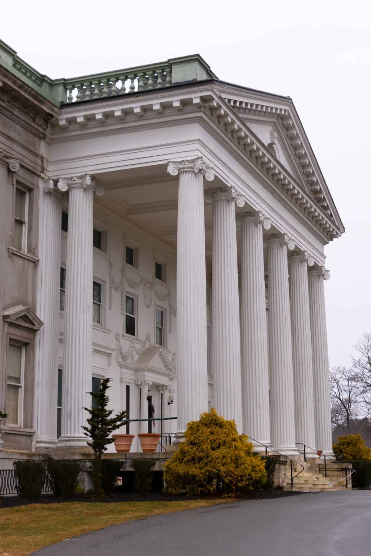 Large neoclassical building with tall white columns, a triangular pediment, multiple windows, and a small staircase leading to the entrance. Bushes and a paved walkway are in the foreground.