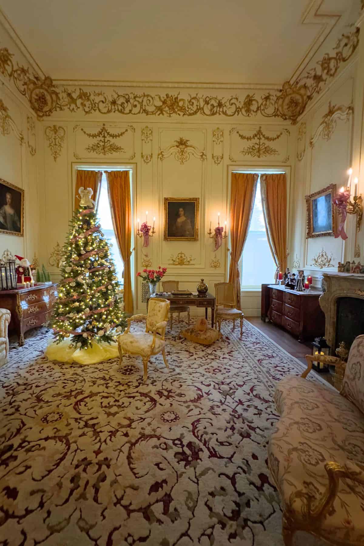 A decorated Christmas tree stands in an ornate, gold-trimmed living room with vintage furniture, patterned carpet, and tall draped windows.