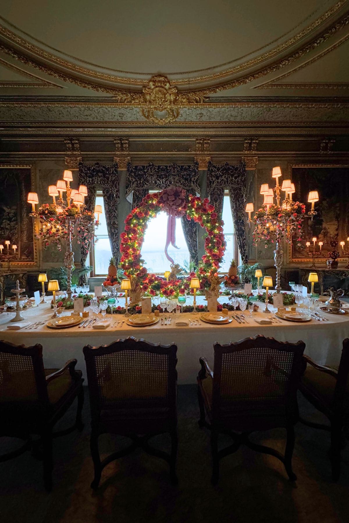 An ornate dining room features a long table set for a meal, elaborate floral wreath centerpiece, and multiple candelabras under decorative ceiling molding.