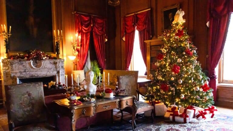 A decorated Christmas tree with presents sits in an elegant, wood-paneled room featuring red curtains, antique furniture, and a marble fireplace adorned with festive ornaments.
