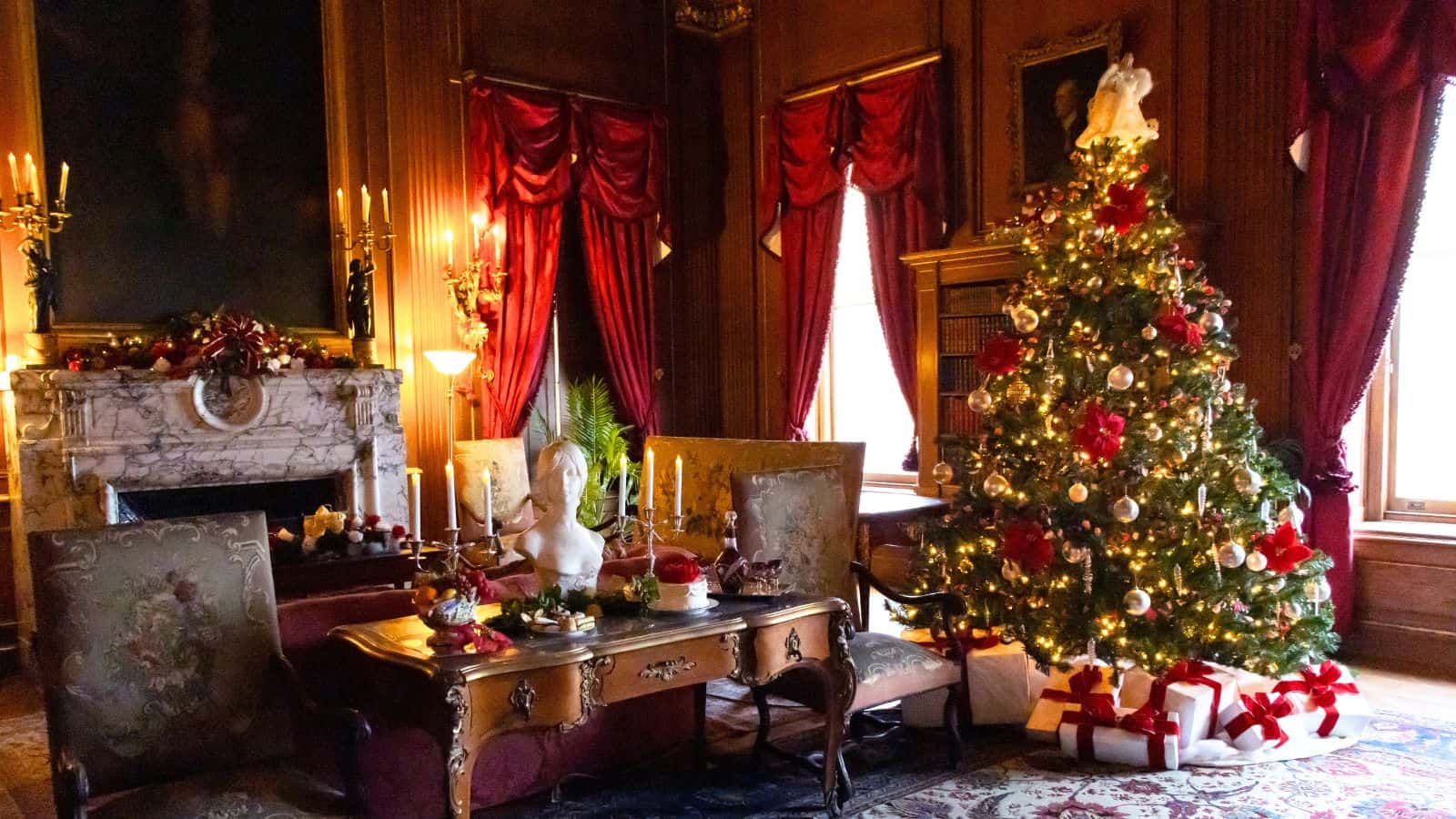 A decorated Christmas tree with presents sits in an elegant, wood-paneled room featuring red curtains, antique furniture, and a marble fireplace adorned with festive ornaments.