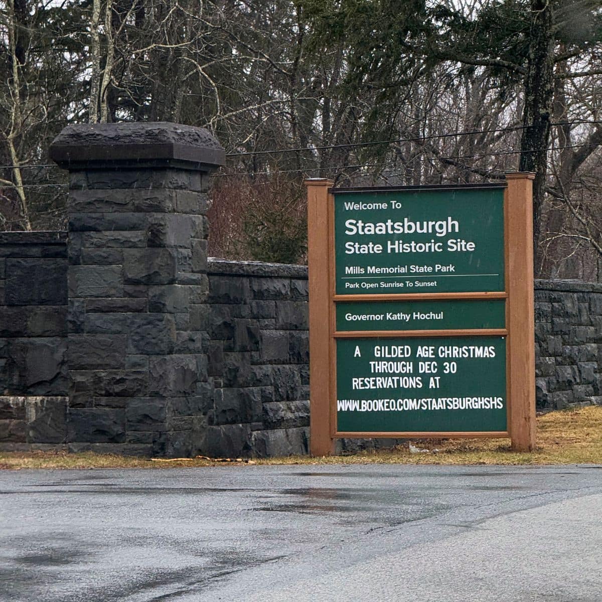 A sign at the entrance of Staatsburgh State Historic Site displays park information, hours, and event details near a stone wall on a cloudy day.