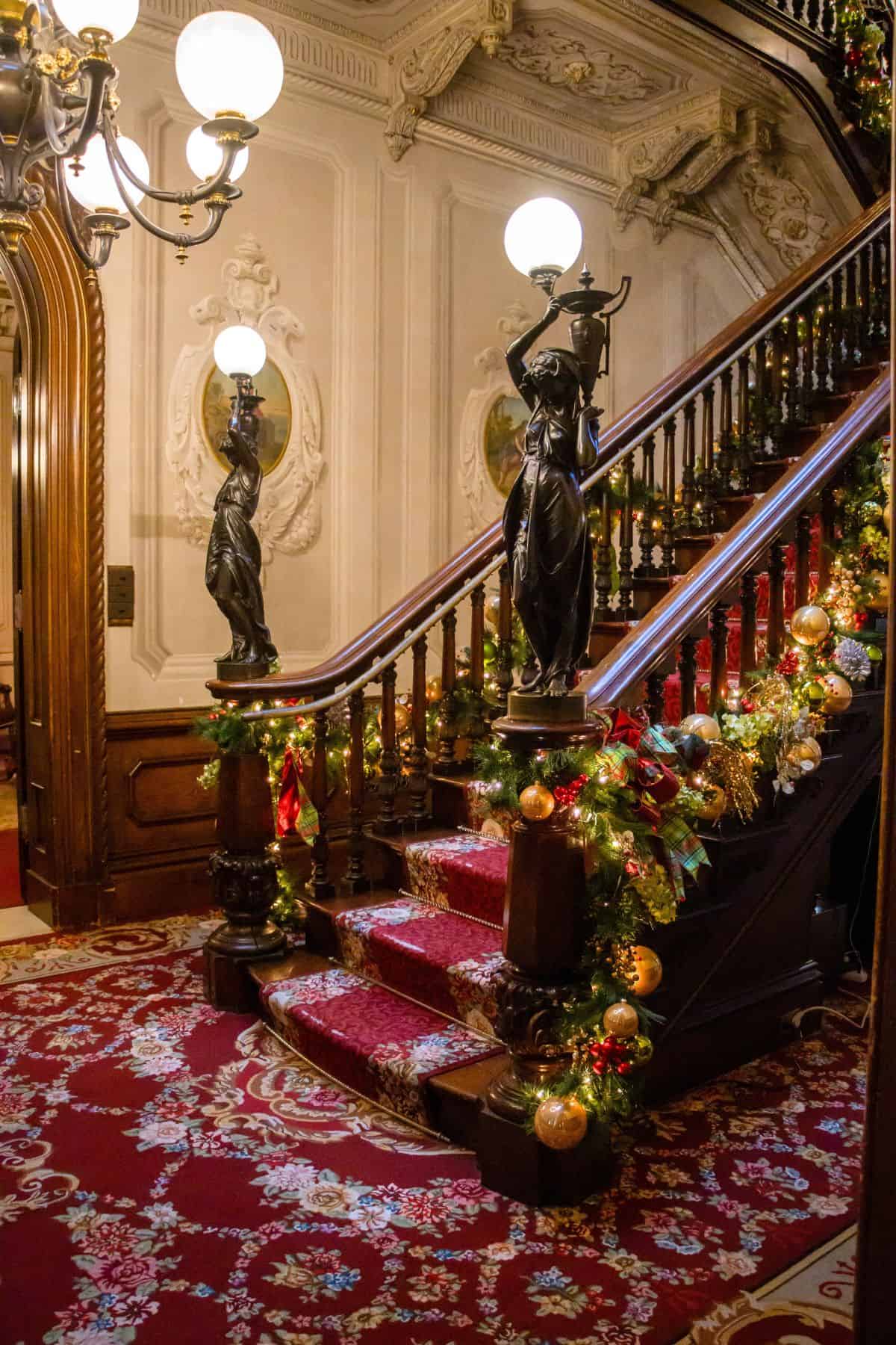 A grand staircase with ornate banisters, red carpet, statues holding lamps, and holiday garlands decorated with gold ornaments.