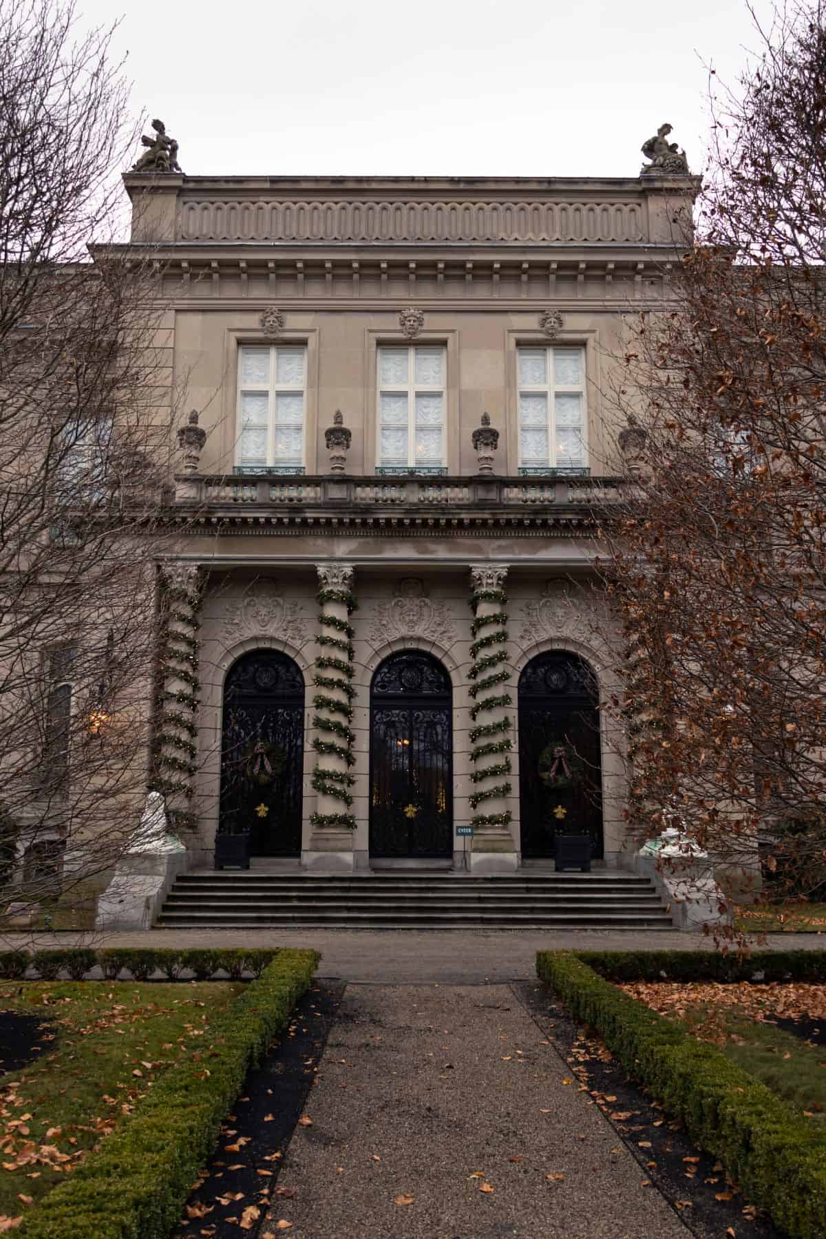 A stately building with arched black doors, tall windows, and garlands wrapping the columns, framed by bare trees and a path lined with trimmed hedges.