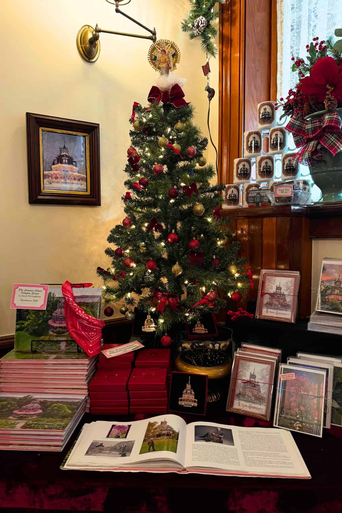 A small decorated Christmas tree is surrounded by stacks of books, framed photos, greeting cards, and mugs, all featuring the same historic building.
