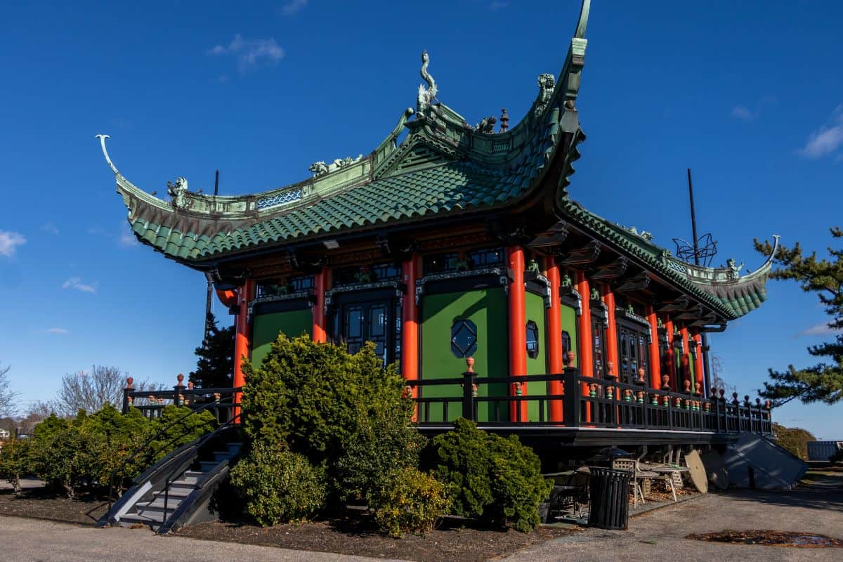 A traditional-style Chinese pavilion with green roof tiles and red columns stands amid shrubs under a clear blue sky.
