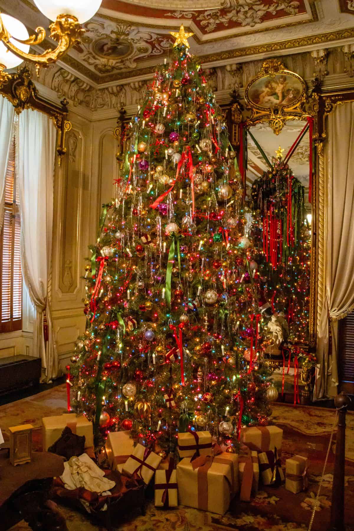 A large, decorated Christmas tree with ornaments and lights stands in an ornate room with wrapped presents underneath and a mirror reflecting the tree in the background.