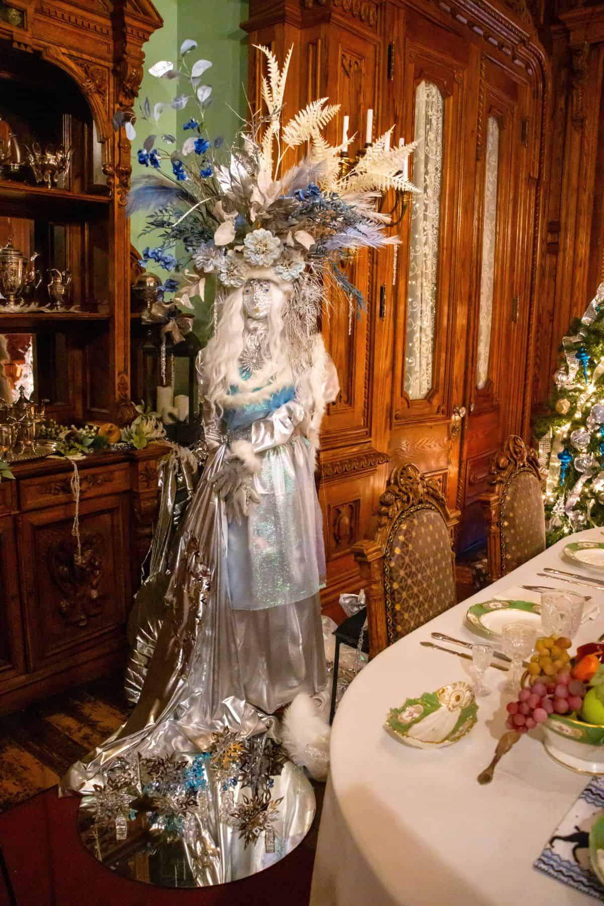 A person dressed in a silver and white costume with a large floral headpiece stands beside a dining table set with dishes, fruit, and ornate wooden furniture.