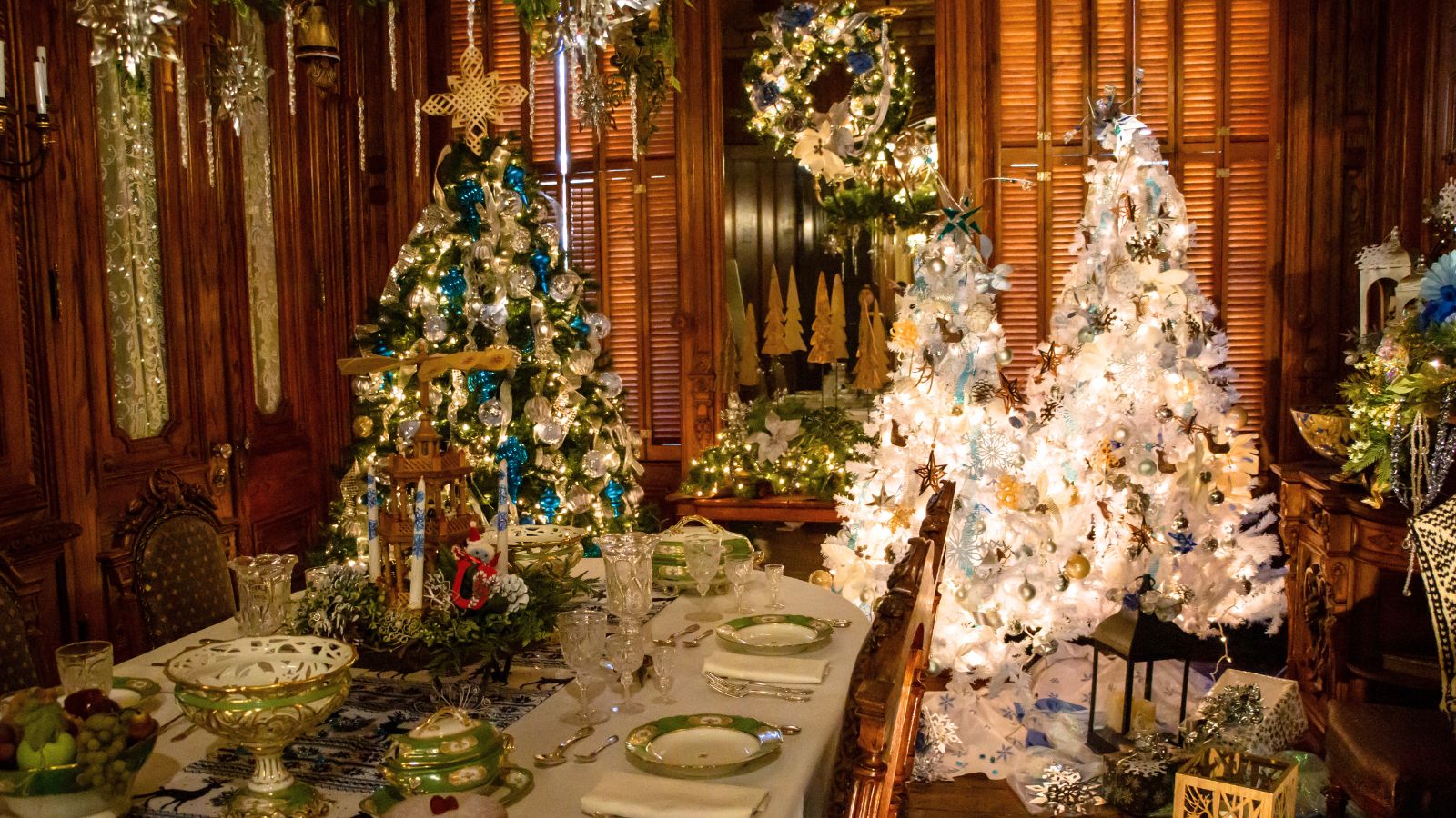 A dining room decorated for Christmas with a set table, ornate chairs, and two elaborately adorned Christmas trees with white and blue ornaments and lights.