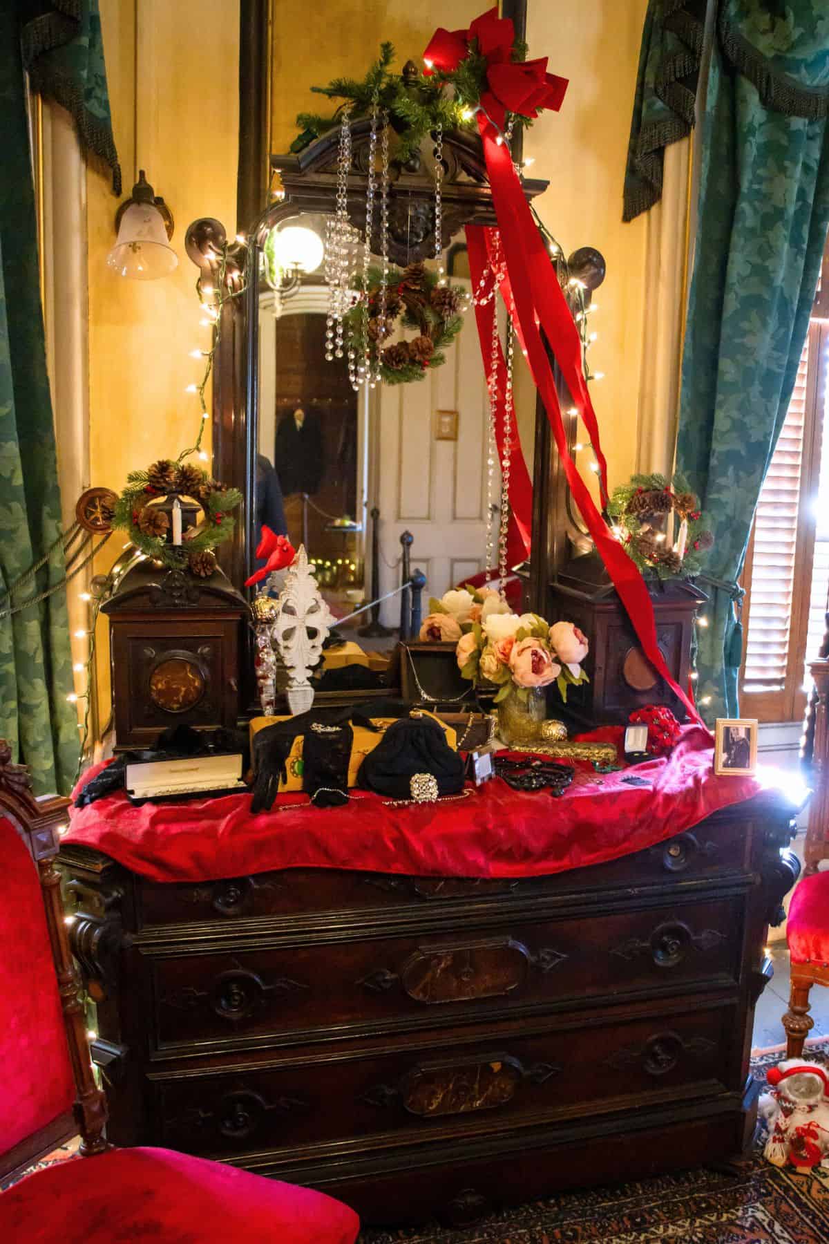 A vintage wooden dresser with a red velvet cover is decorated with flowers, jewelry, a mask, wreaths, lights, and a large red ribbon, in a room with green curtains and antique furniture.