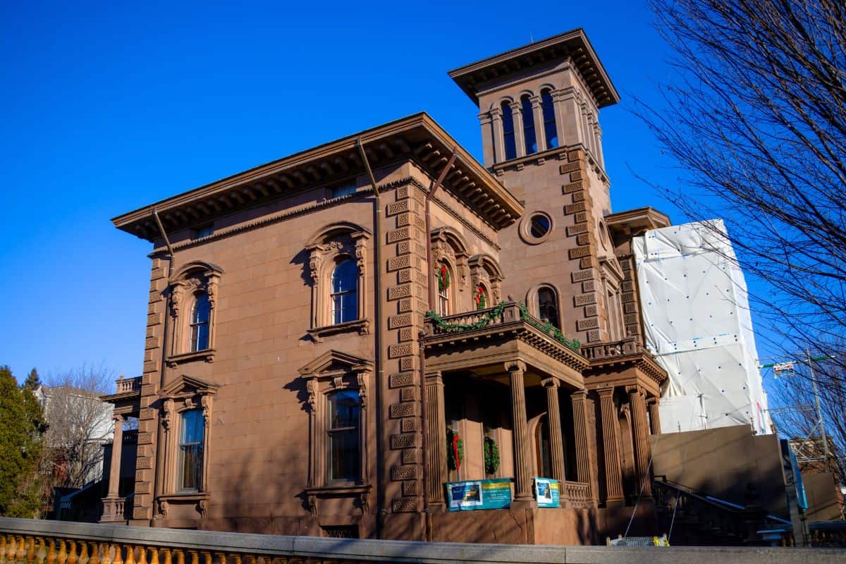 A brown historic mansion with arched windows, ornate details, and a tall central tower, set against a clear blue sky with some trees and construction nearby.