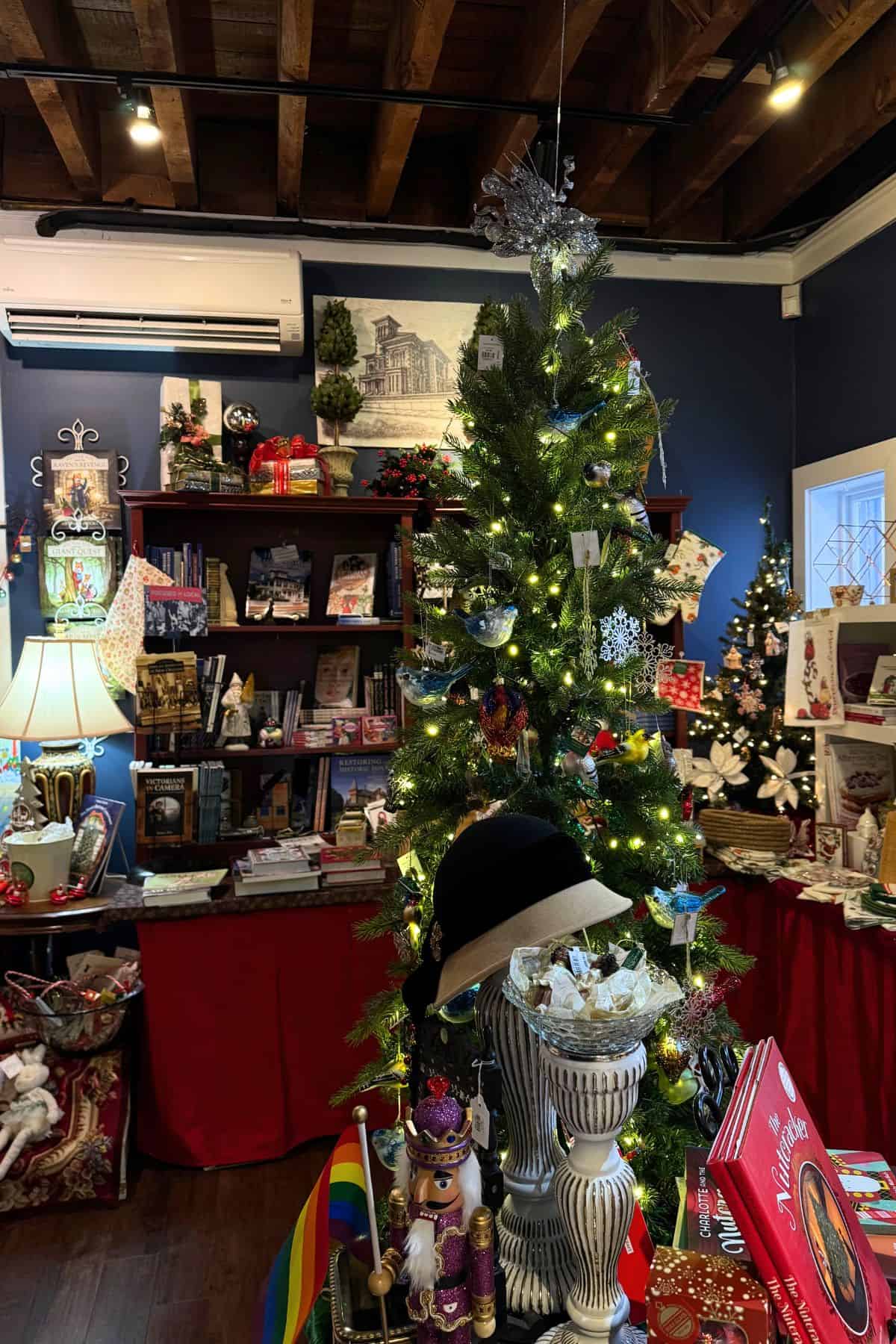 A decorated Christmas tree with ornaments and lights stands in a festive shop, surrounded by holiday-themed gifts and decorations, evoking the charm of Christmas at Victoria Mansion.