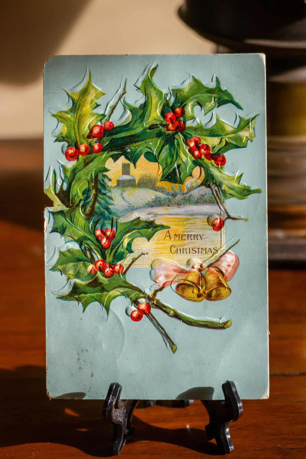 A vintage postcard with holly leaves, red berries, gold bells, and the text "A Merry Christmas," displayed upright on a stand.