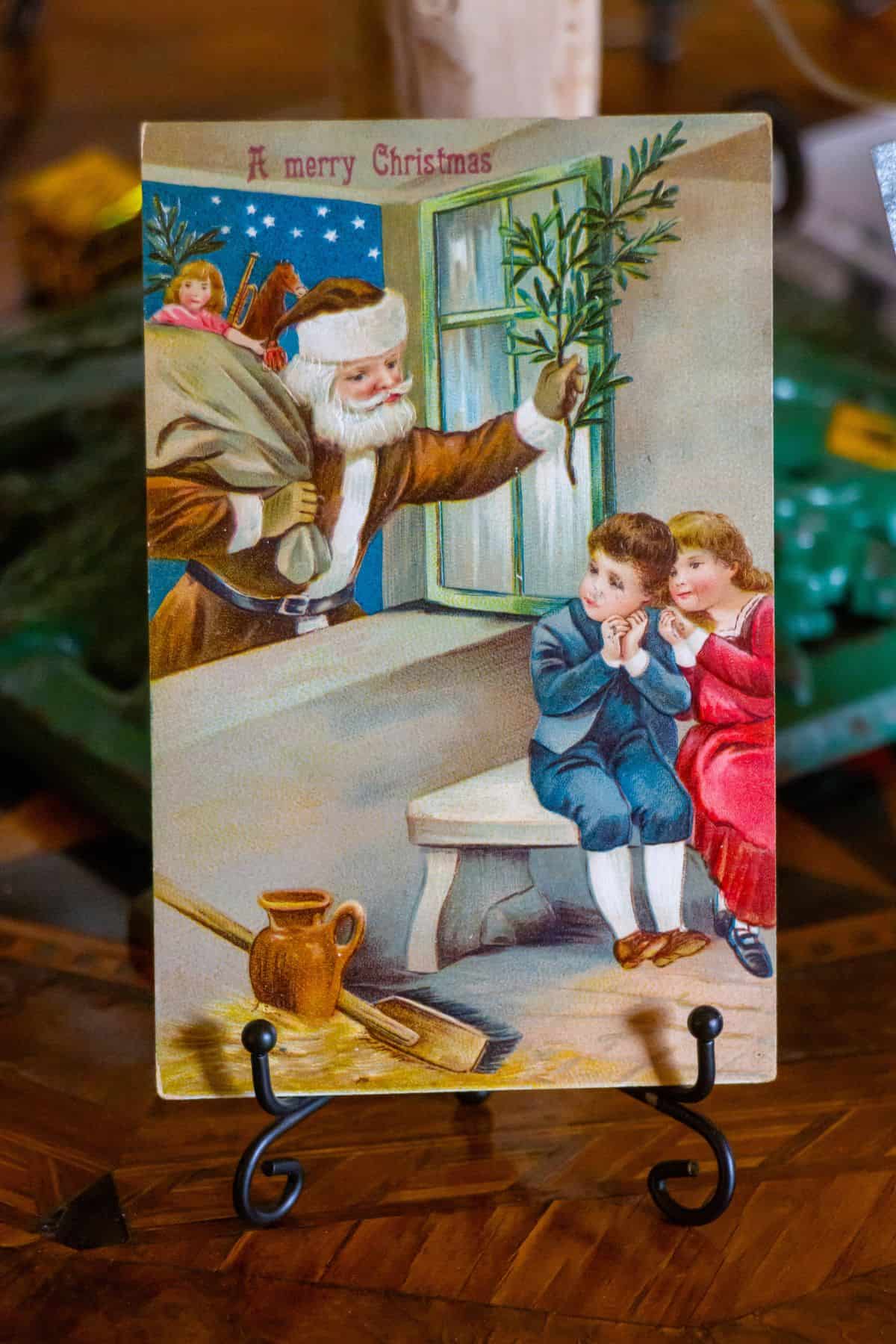 Vintage Christmas card showing Santa Claus holding a bag and greenery by a window, with two children sitting on a bench indoors, one in blue and one in red, looking at him.