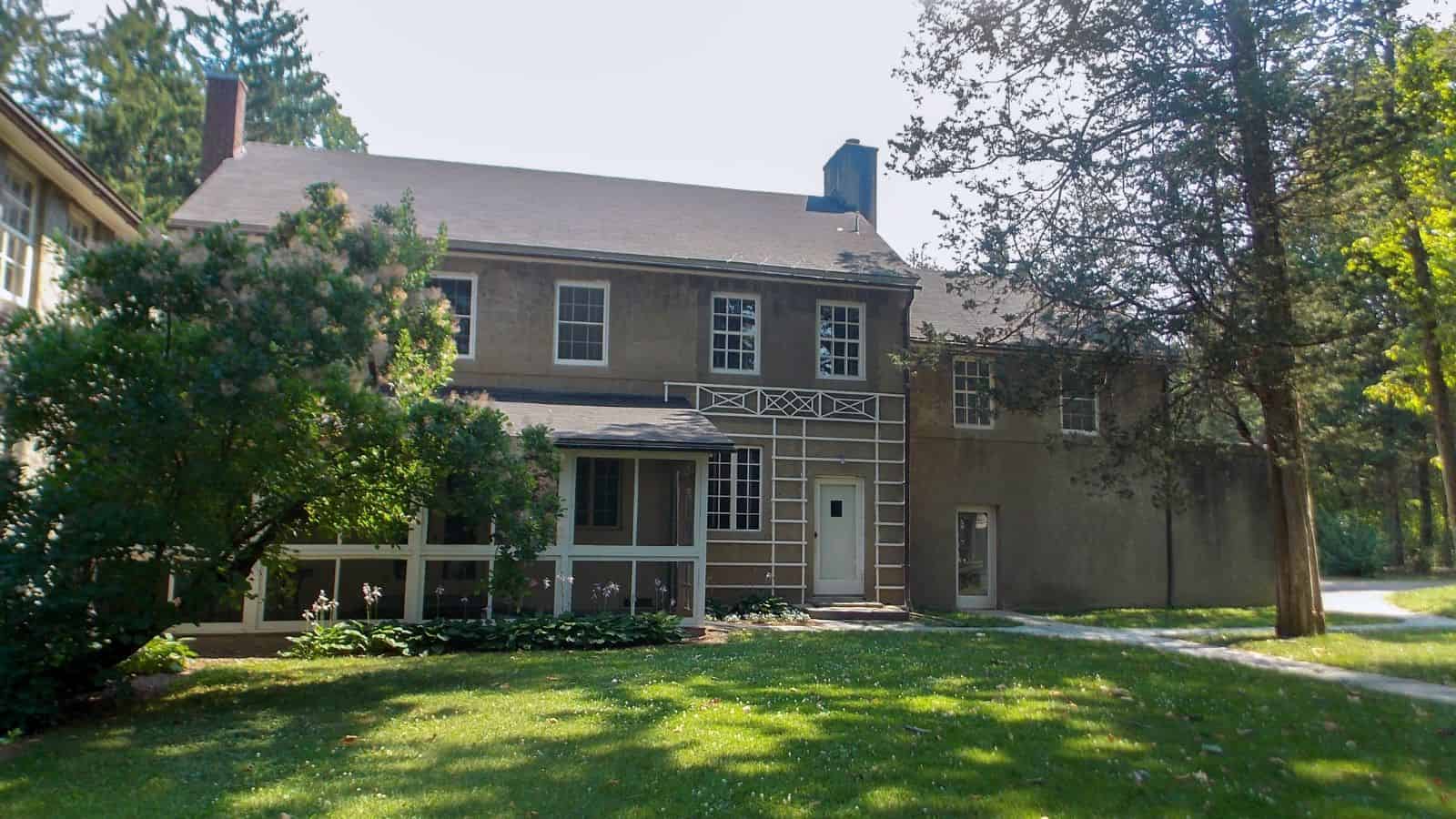 Two-story house with a screened porch, beige exterior, and a small balcony, surrounded by green lawn and trees on a sunny day.