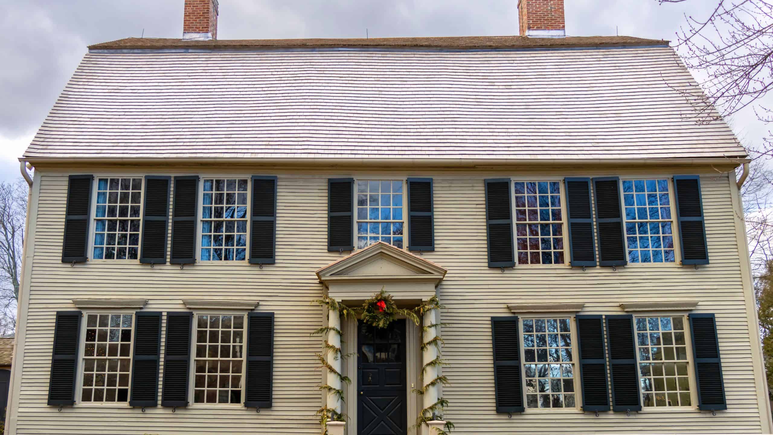 A colonial-style house with cream-colored siding, black shutters, a central door with a decorative garland, and two brick chimneys, under a cloudy sky.