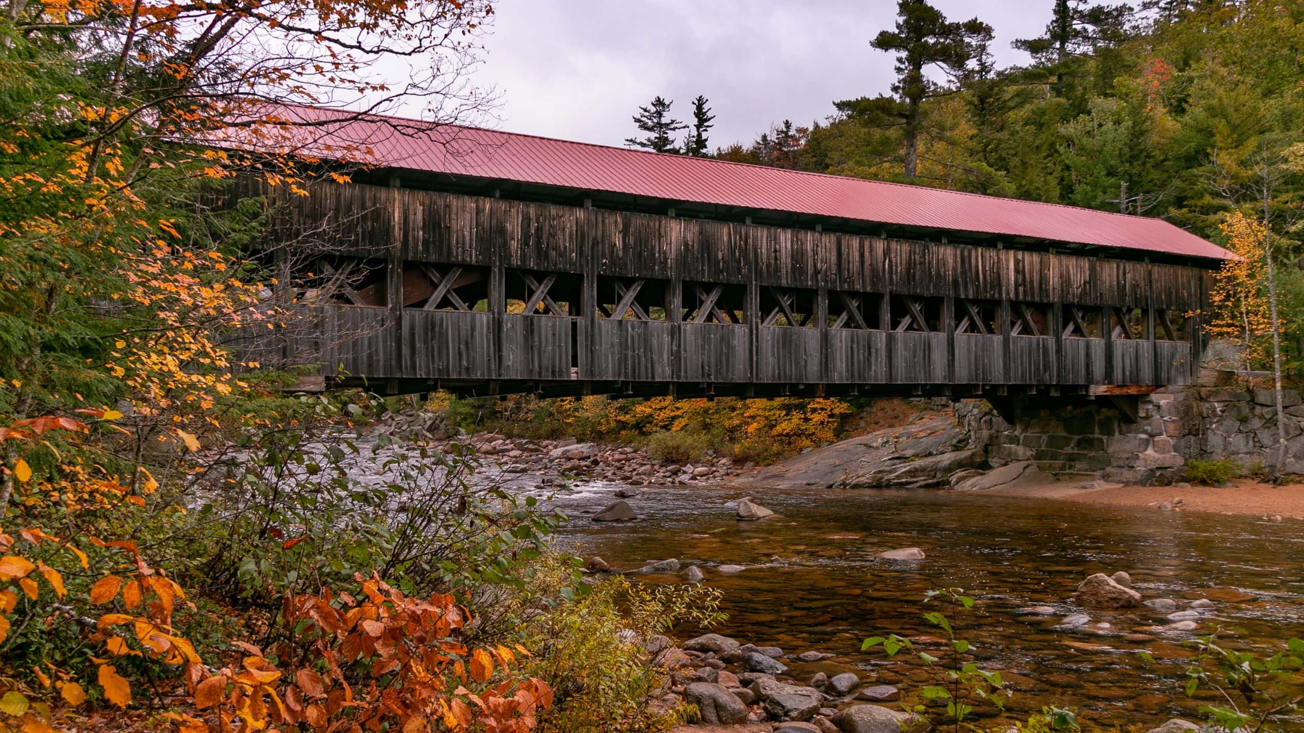 A long wooden covered bridge with a red roof spans over a shallow rocky river, surrounded by autumn trees and foliage.