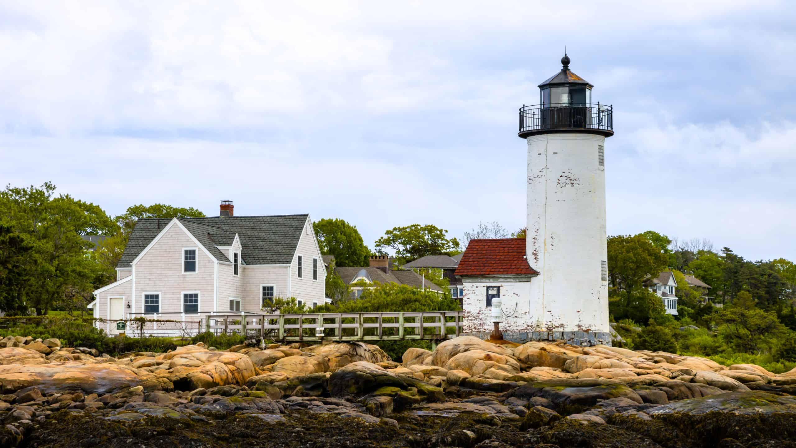 A white lighthouse with a black top, part of the iconic lighthouses in Massachusetts, stands next to a white house, surrounded by trees and rocks under a cloudy sky.