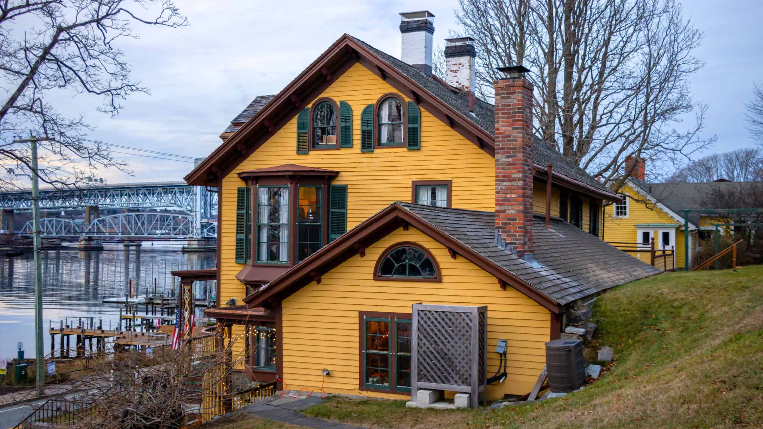 A yellow wooden house with green shutters and multiple chimneys is situated on a grassy slope by a riverside, with a bridge visible in the background.