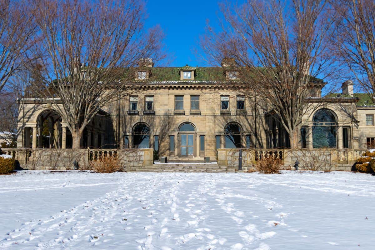 Large stone mansion with arched windows and columns, surrounded by leafless trees, sits behind a snow-covered lawn with visible footprints under a clear blue sky.