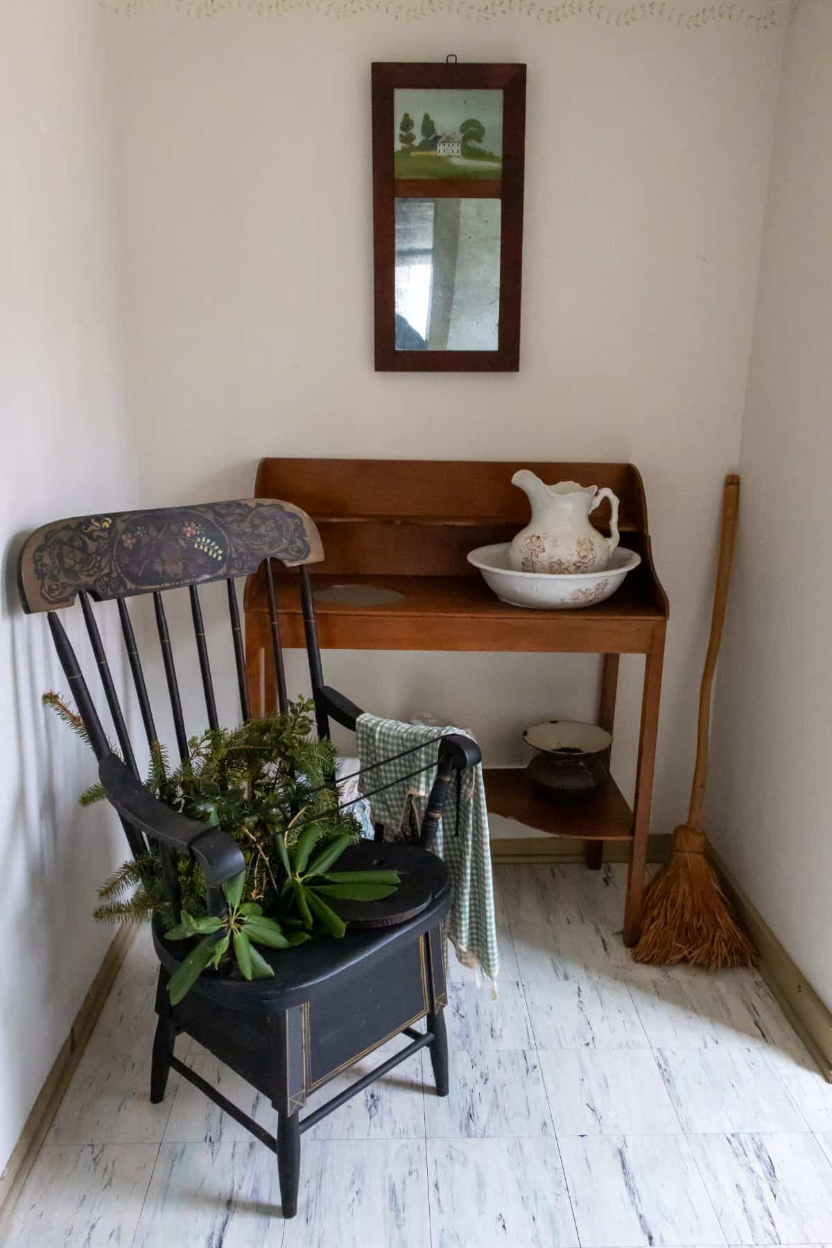 A wooden chair with greenery, a green-checkered cloth, a wooden table with a pitcher and bowl, a broom, and a framed picture in a small corner room.