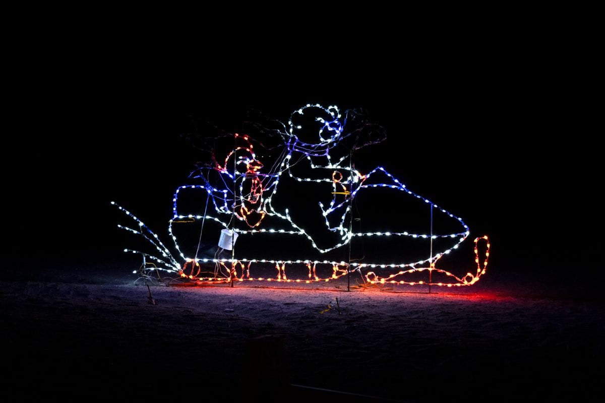 Holiday light display featuring two figures riding a snowmobile, outlined with red, white, and blue lights against a dark background.