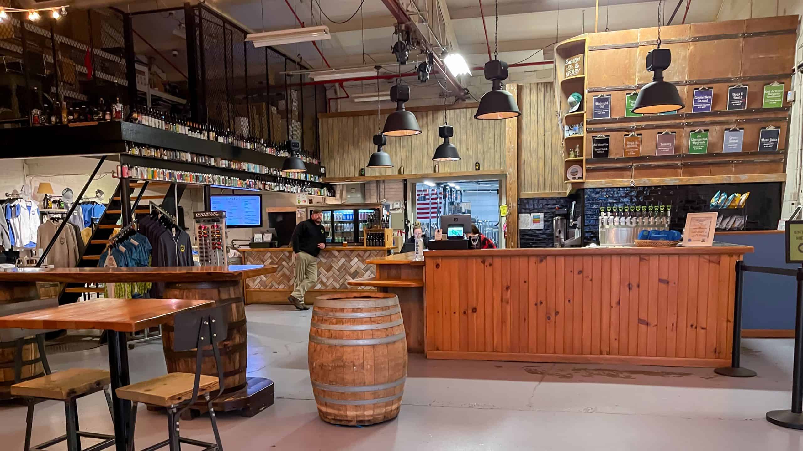 Interior of a brewery taproom with wooden bar, beer taps, menu boards, tables, chairs, and barrels, with a person standing behind the counter.