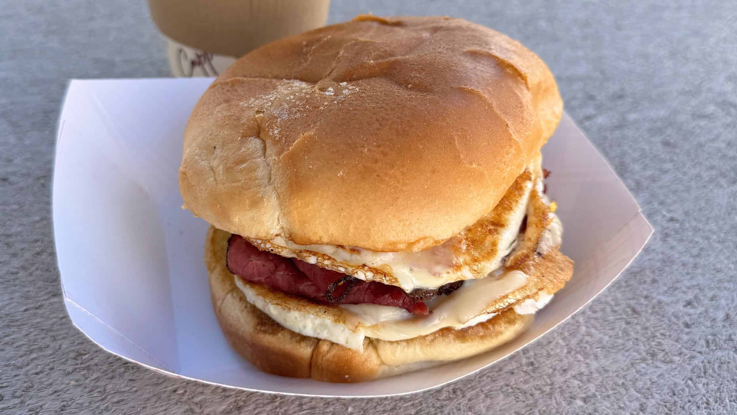 A breakfast sandwich with fried eggs, meat, and melted cheese in a soft bun, served in a paper tray with a cup in the background.