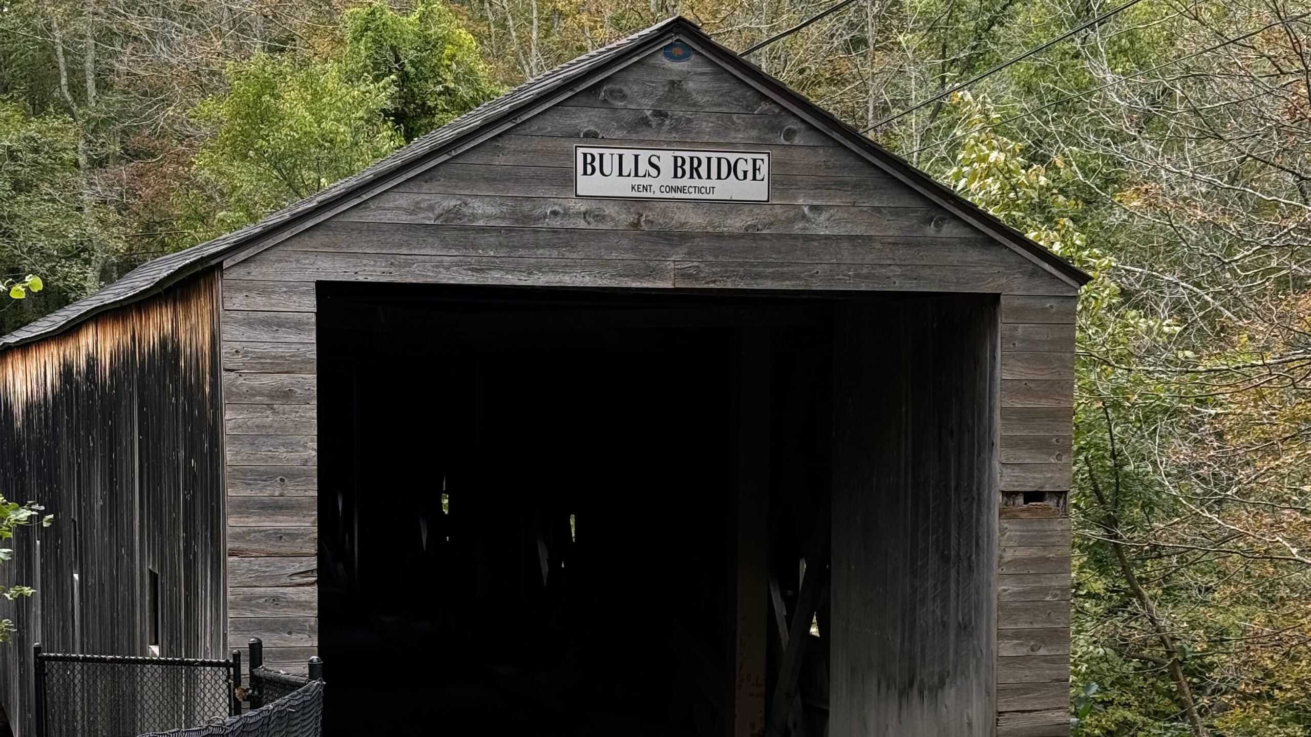 A classic New England covered bridge labeled "Bulls Bridge, Kent, Connecticut," stands surrounded by trees with lush greenery and a black fence in the foreground.