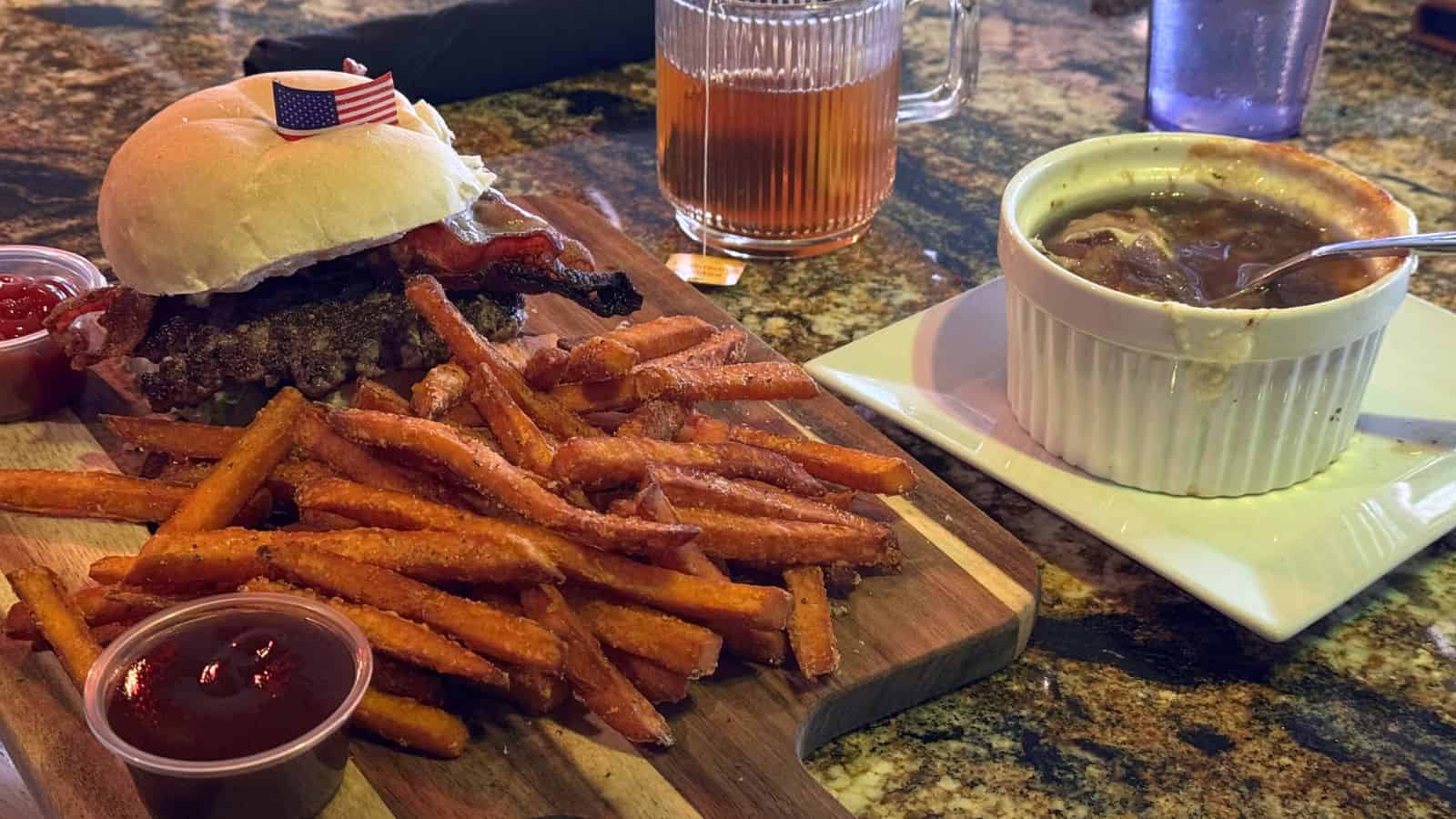 Burger with bacon, sweet potato fries, ketchup, and a bowl of French onion soup with a spoon, served on a wooden board and plate—delicious food in Niantic, Connecticut on a cozy restaurant table.