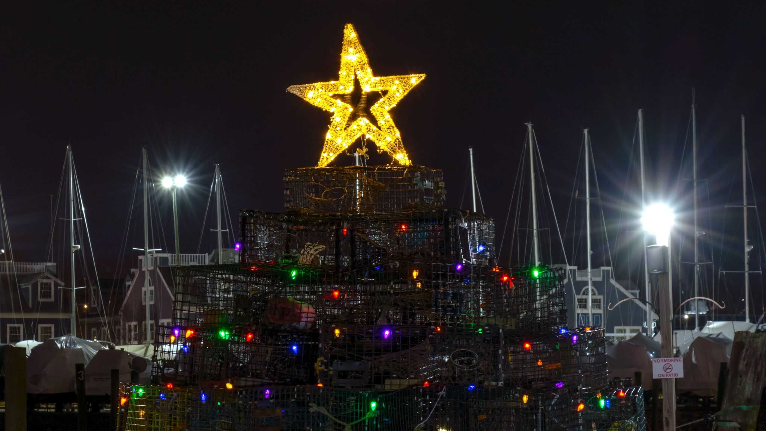 A Christmas tree made of stacked lobster traps decorated with colorful lights and topped with a glowing star, with boats and masts visible in the background at night.